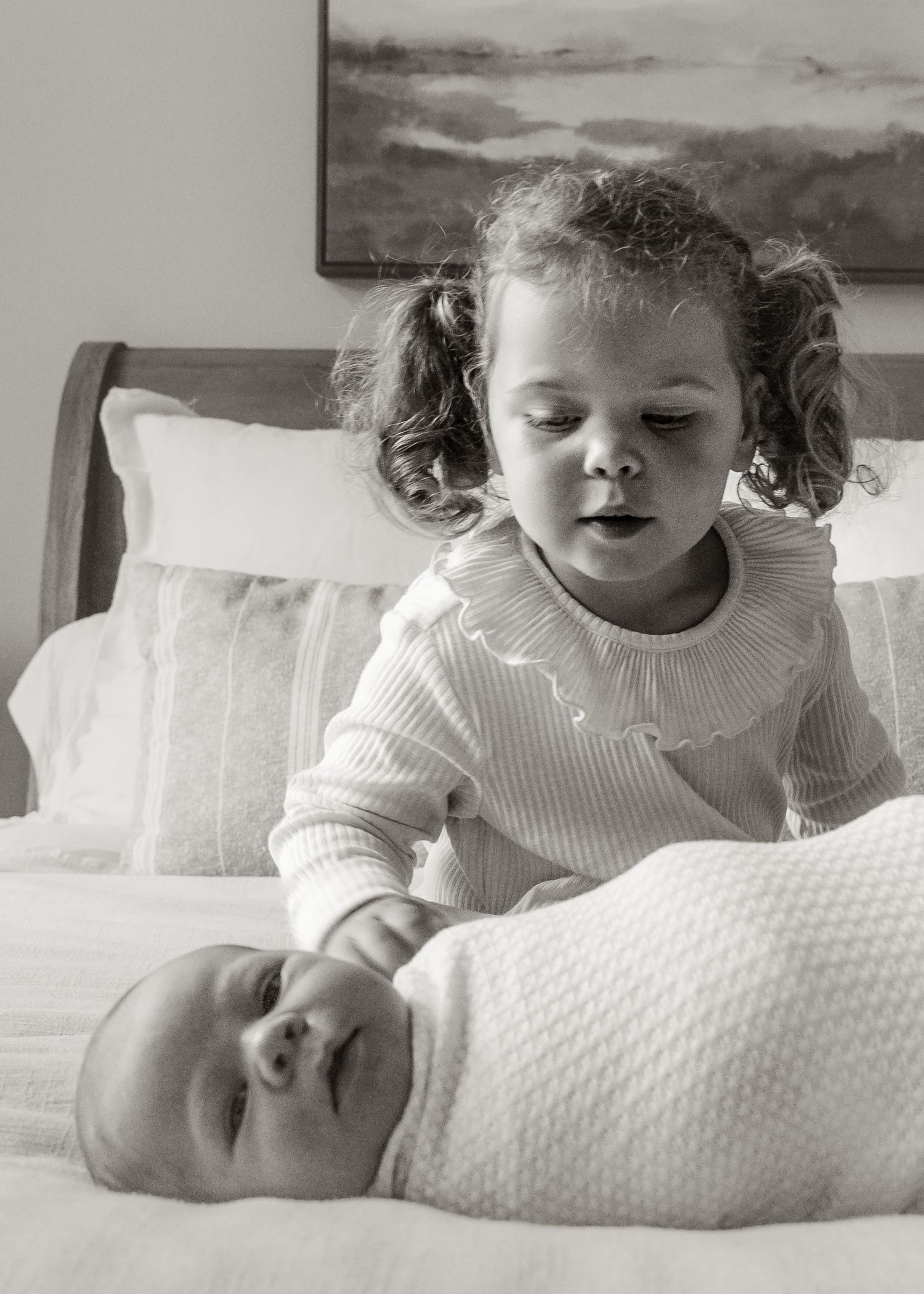A tender black and white photograph showing a baby in a white blanket lying on a bed while being watched from above.