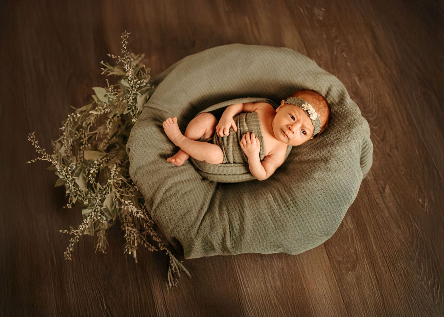 A sleeping newborn baby curled up on a round sage green pillow next to delicate dried flowers on wooden flooring.