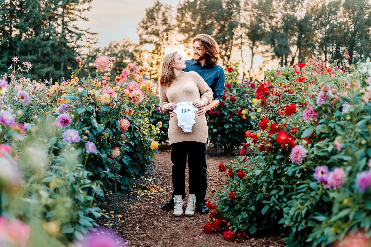 A expecting couple embraces in a garden path surrounded by colorful dahlias and flowers.