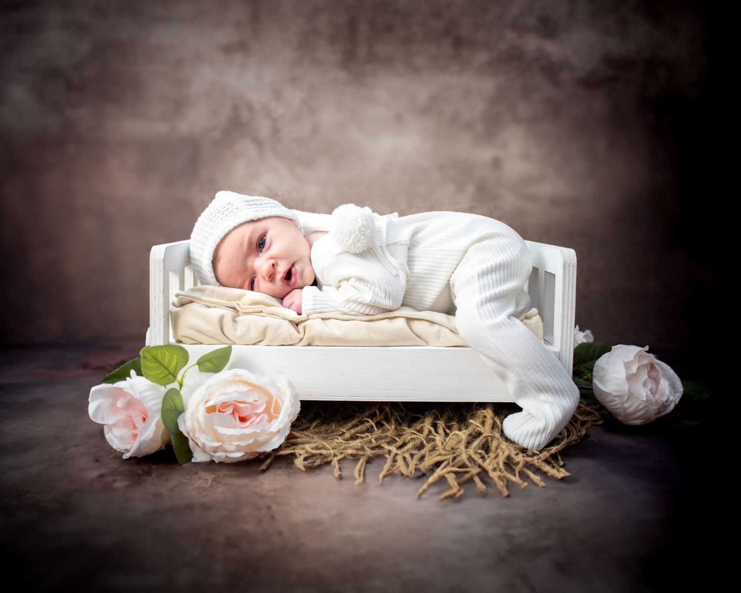 A sleeping newborn in white outfit rests on a miniature bed with soft pink roses and rustic hay backdrop.