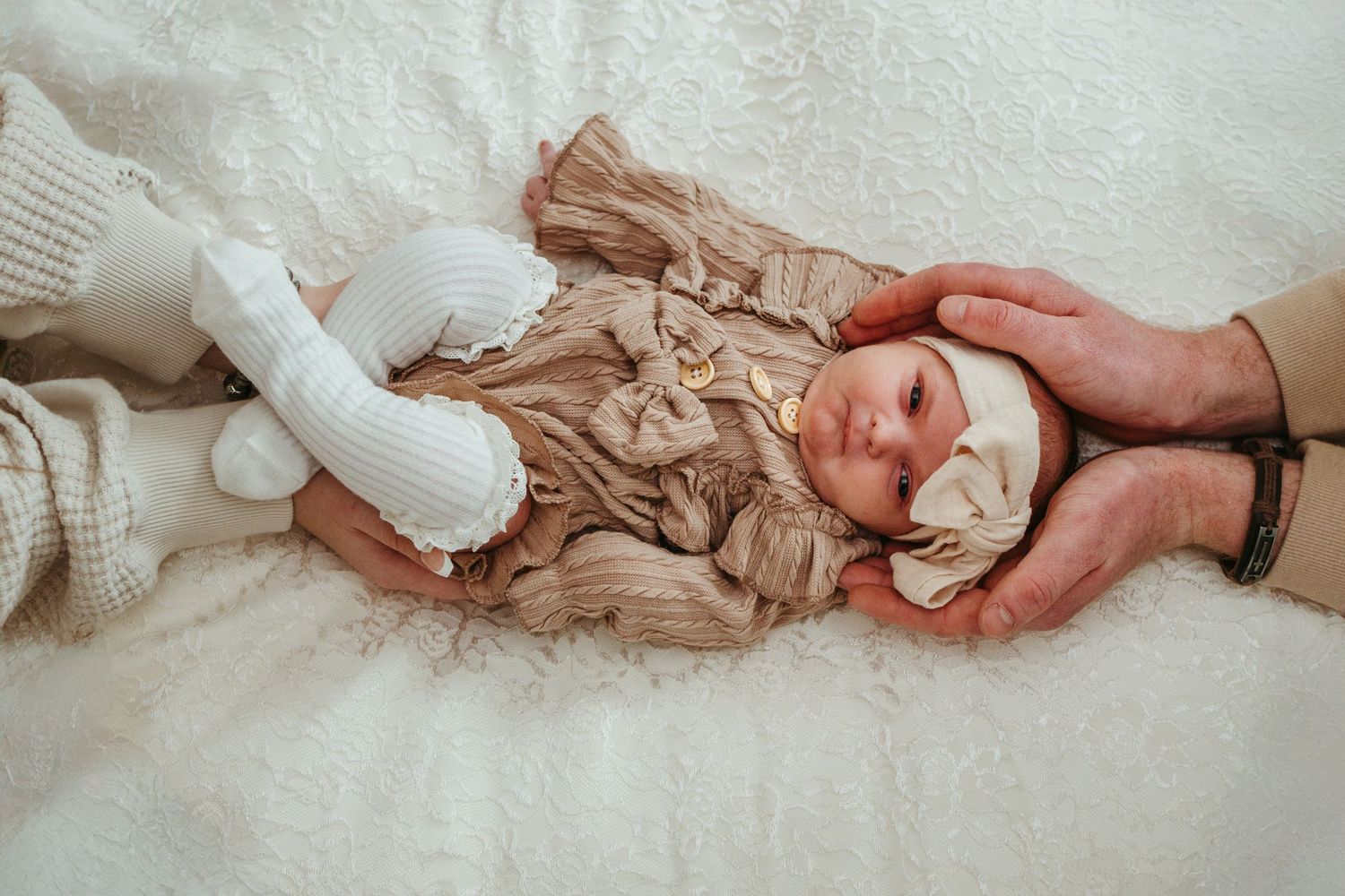 A delicate newborn baby swaddled in white rests gently on a soft neutral-colored blanket with fingers holding it tenderly.