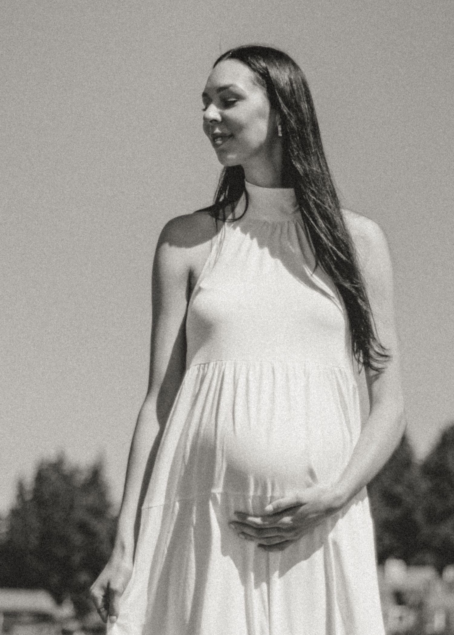 Pregnant woman in white halter dress poses outdoors in black and white photography.