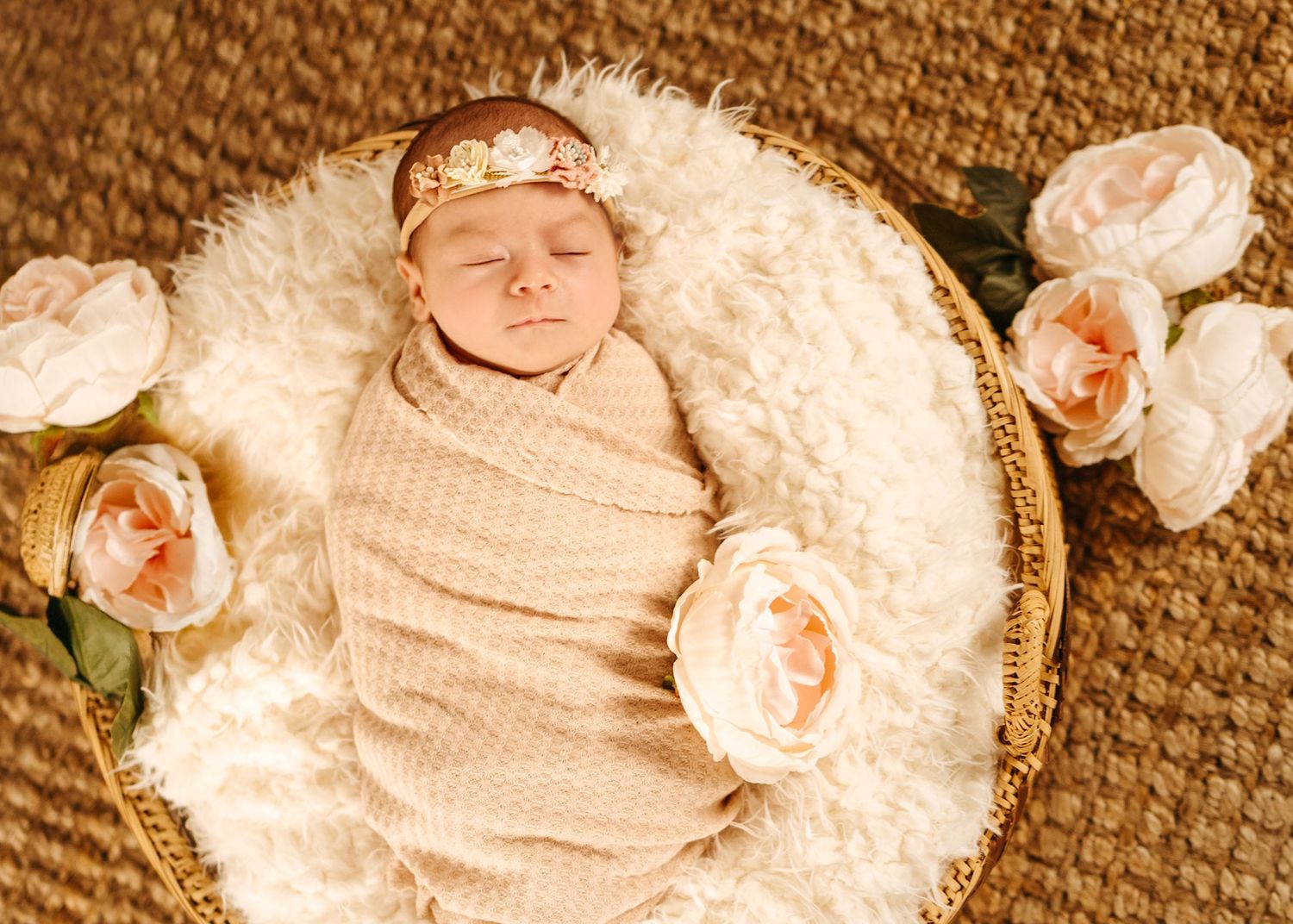 A sleeping baby swaddled in beige fabric lies on white fur surrounded by soft pink roses in a round basket.