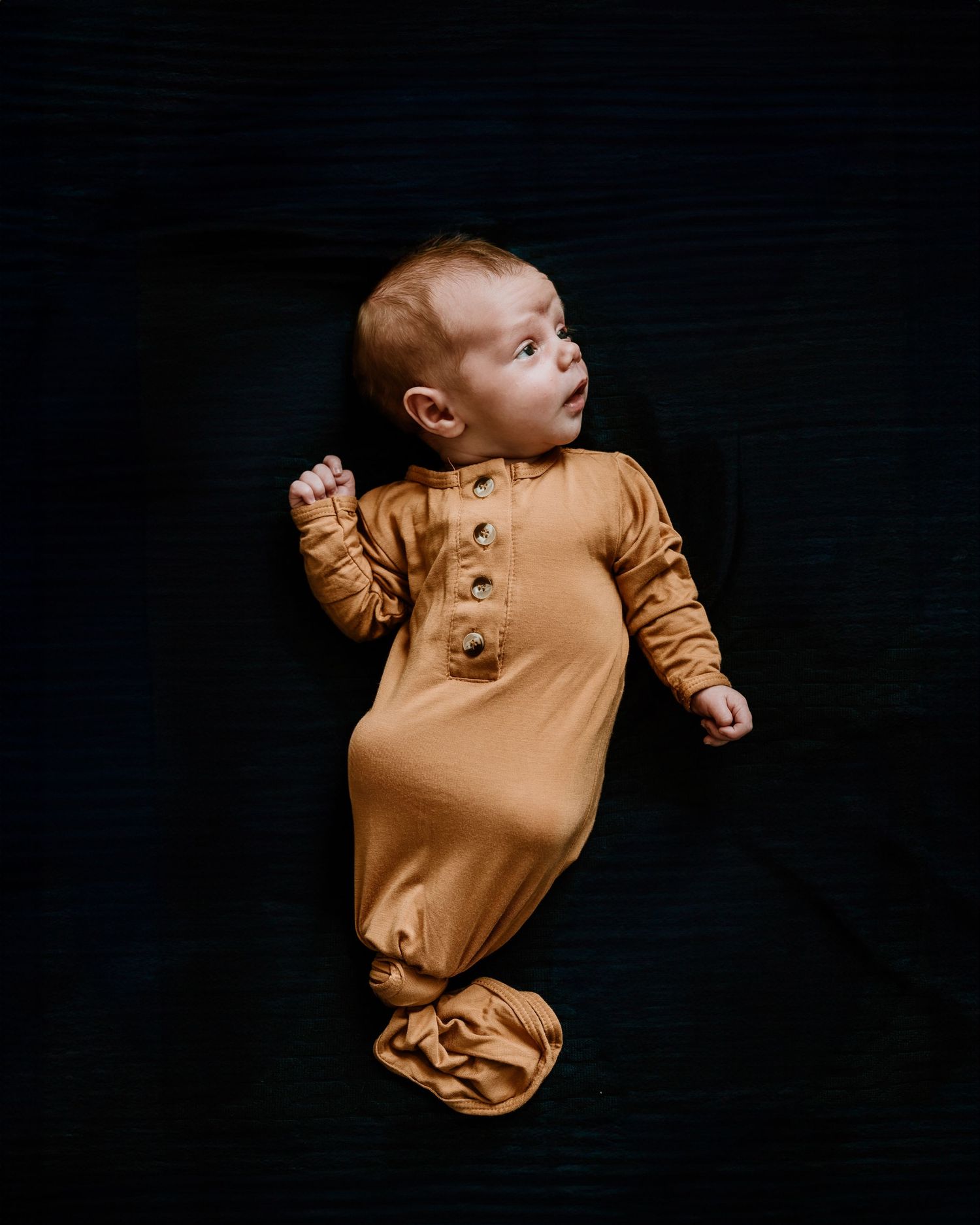 A baby in a caramel-colored henley romper lies against a dark background looking to the side.