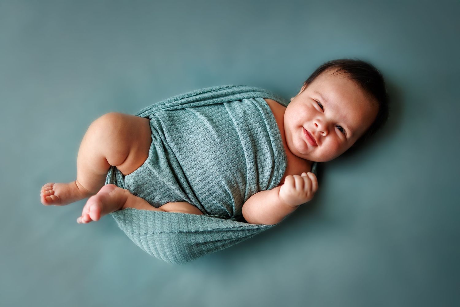 Happy baby in teal romper lies on turquoise blue background during newborn photo session.