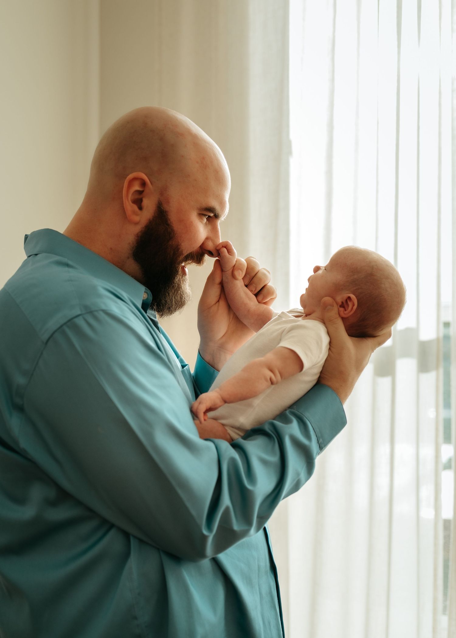 A bearded man in turquoise shirt holds and interacts playfully with a small baby near a window with natural light.