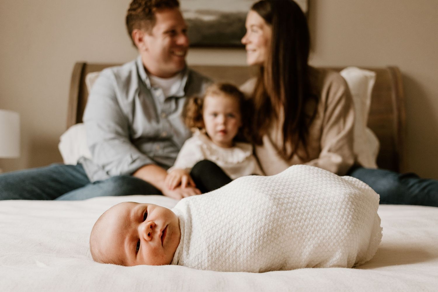 A newborn baby lies swaddled in a white blanket on a bed while a family sits together in the background.