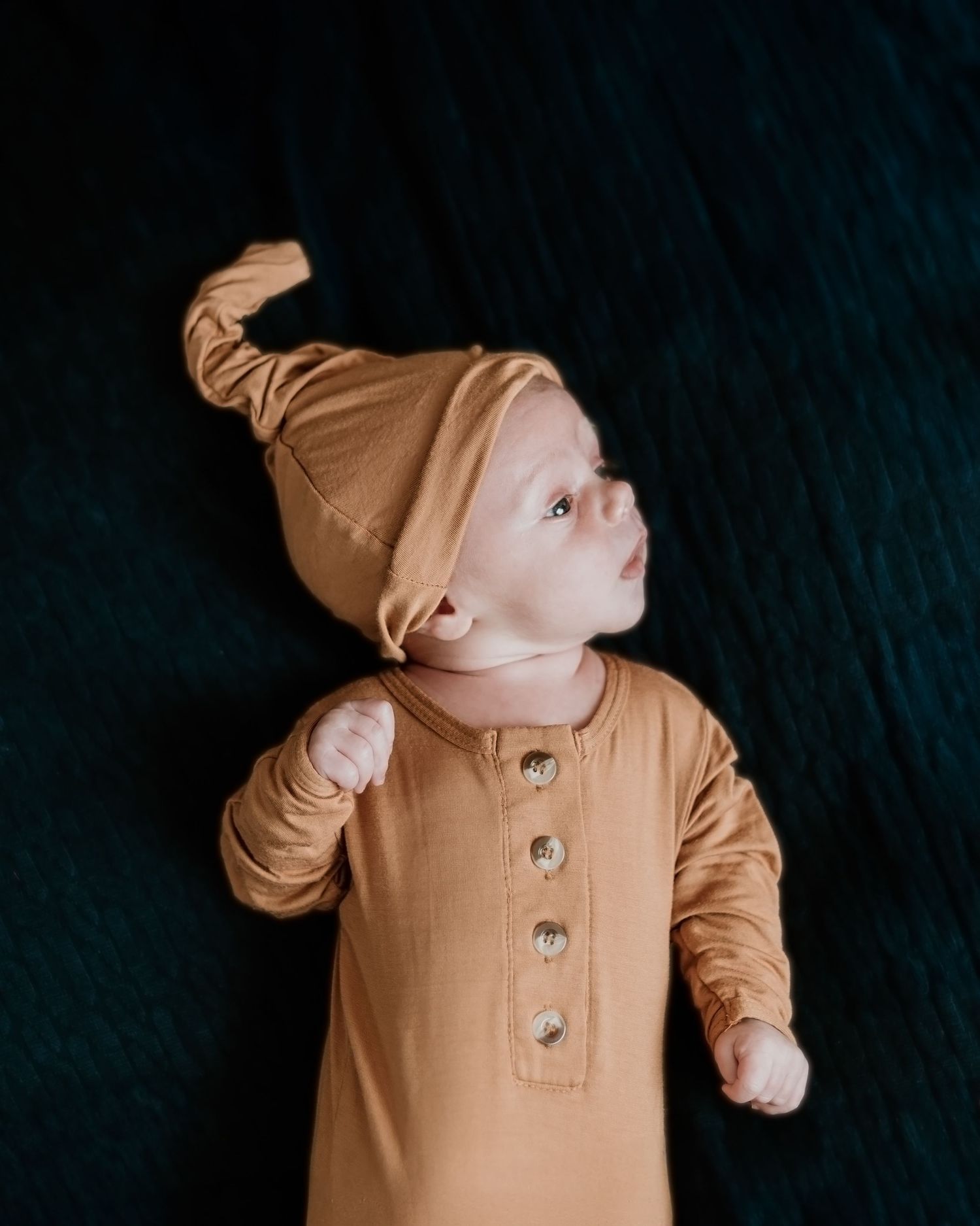 A small child wearing a tan knotted hat and matching button-up romper outfit against a dark background.
