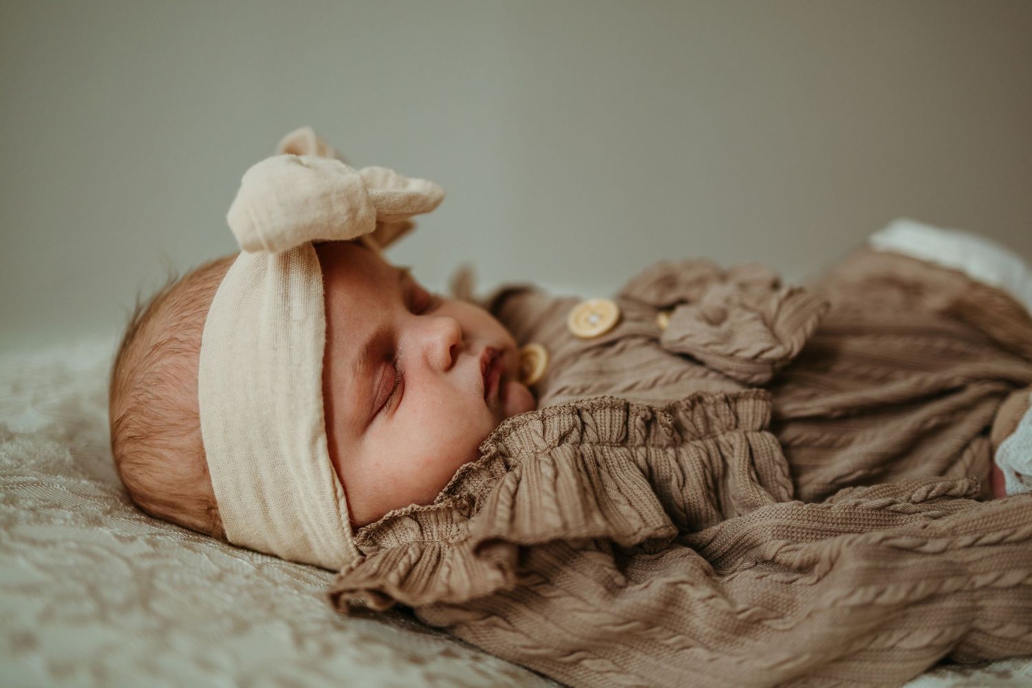 A sleeping newborn baby wearing a cream headband and brown ruffled outfit lies on soft white bedding.