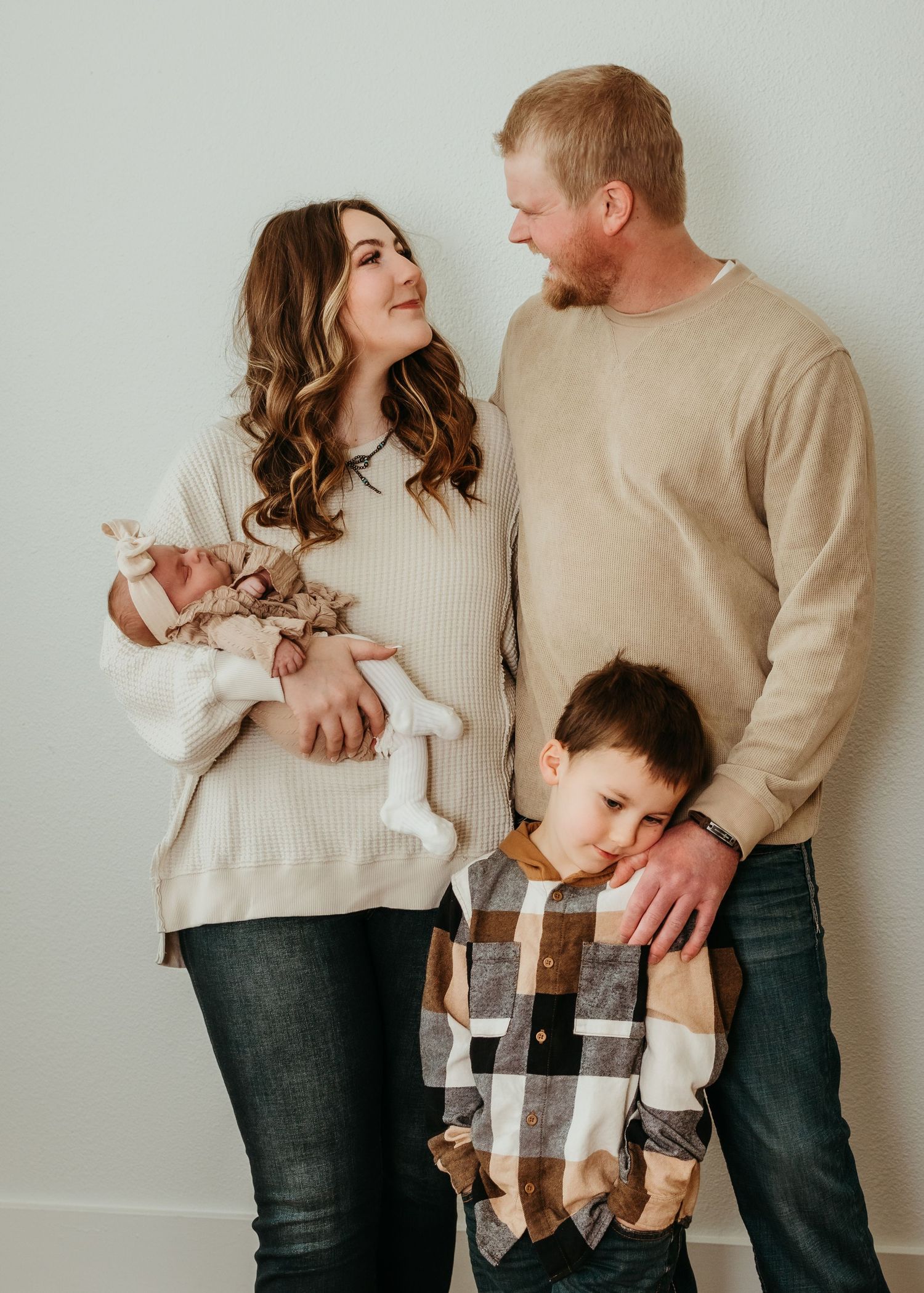 Family wearing coordinated neutral sweaters and plaid posing together against a light gray wall for a heartwarming portrait.