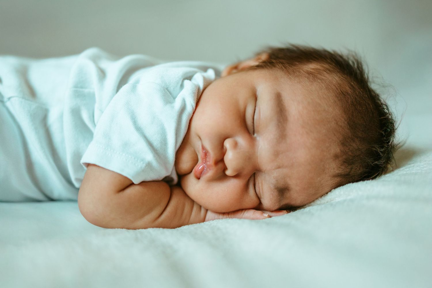 A sleeping baby in a light blue onesie rests peacefully on cozy bedding.