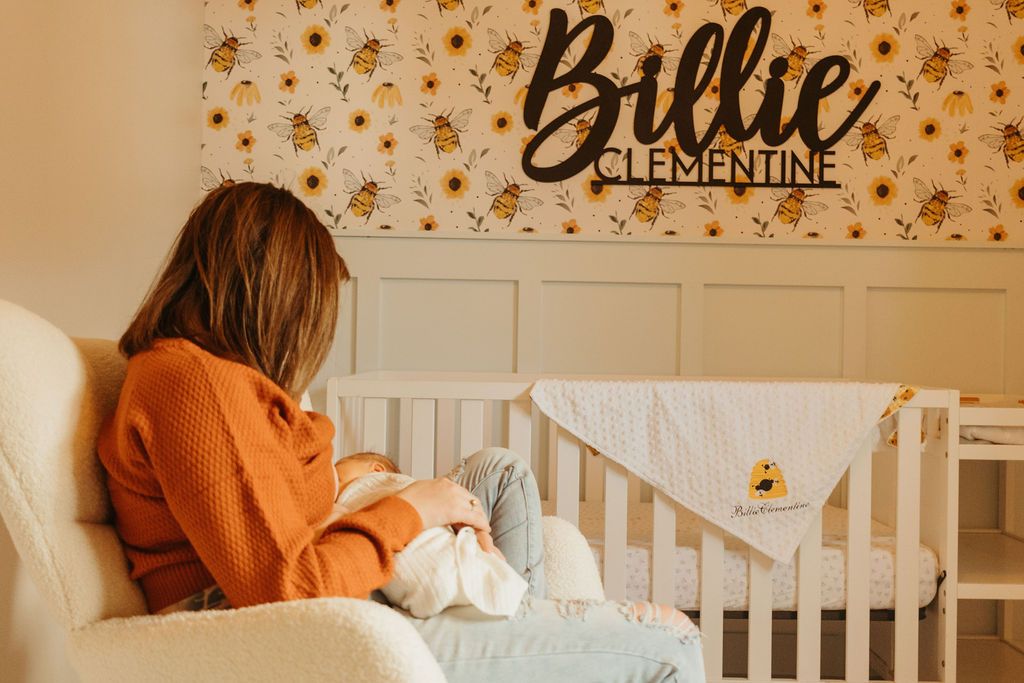 A person in an orange sweater sits in a nursery chair next to a white crib with floral decor on the wall.