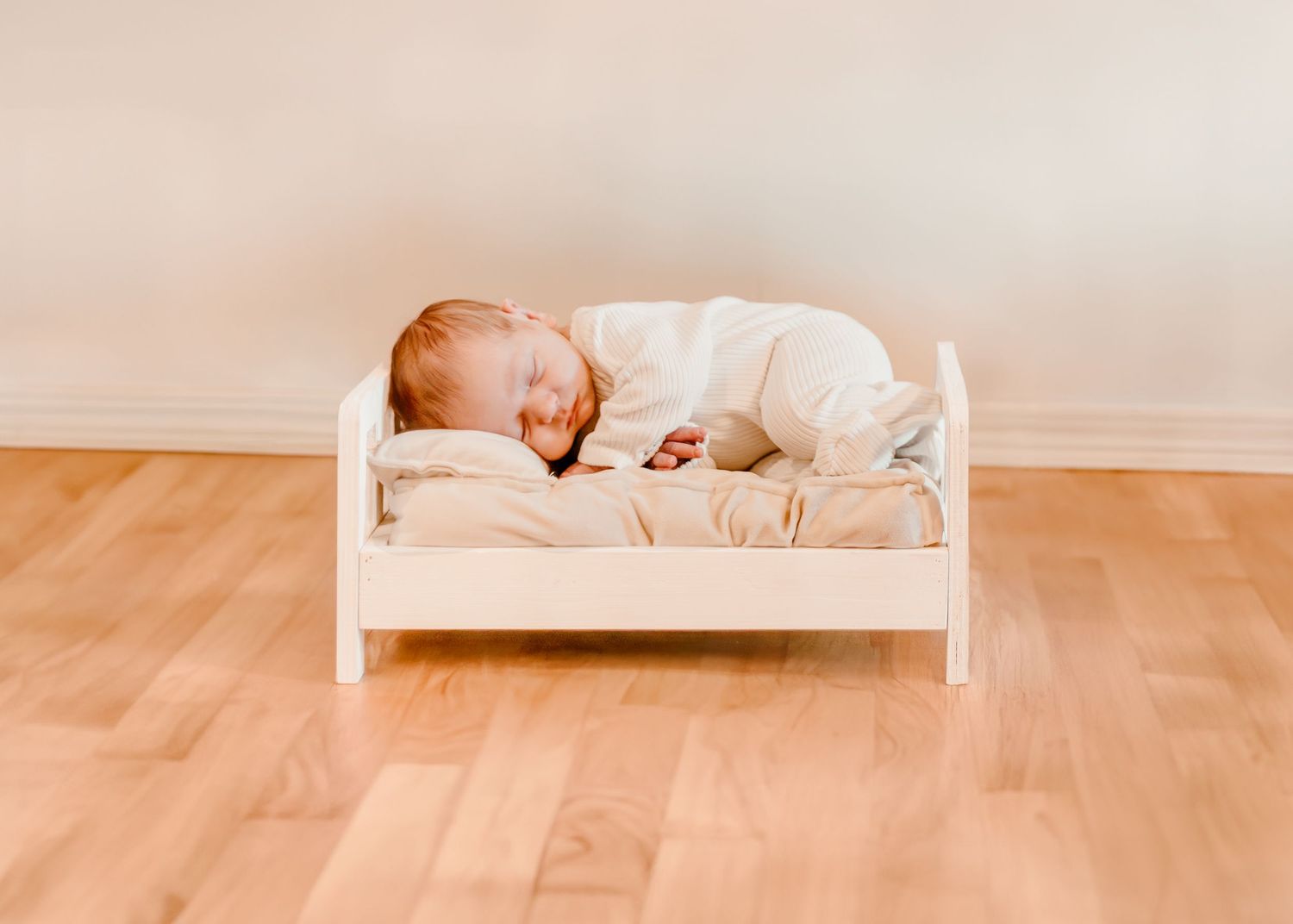 Small child sleeping peacefully in a white wooden doll bed on hardwood floor.