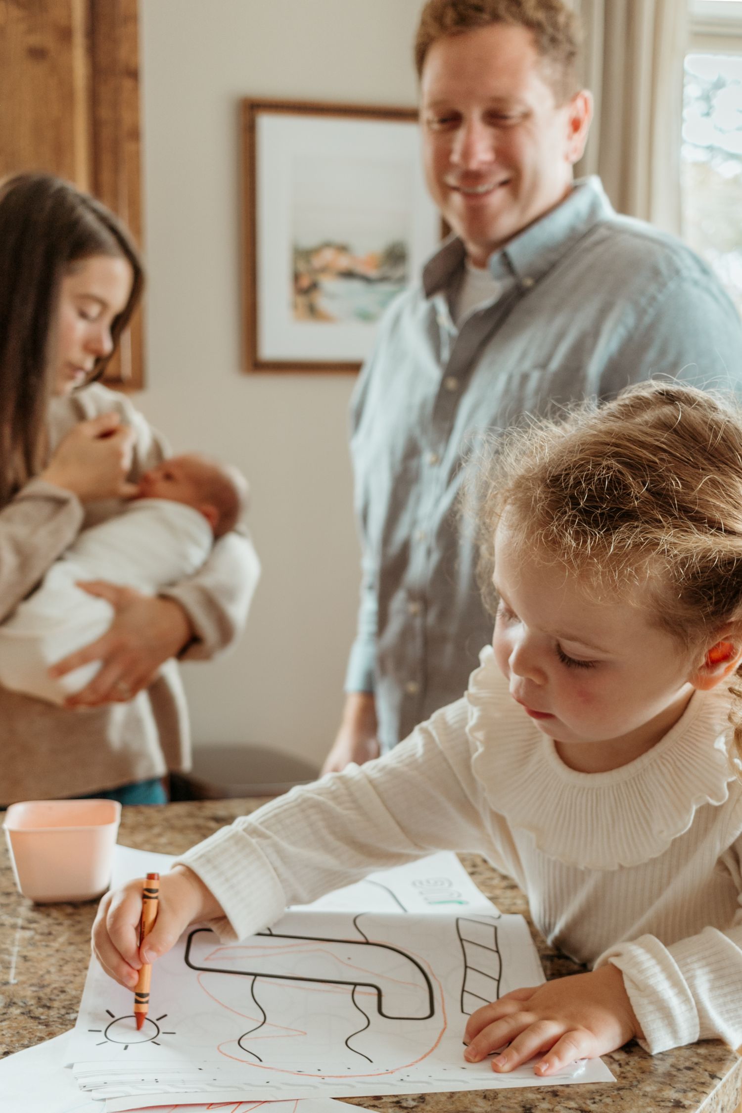 A child draws with crayons at a counter while adults gather around a newborn baby in a cozy home setting.
