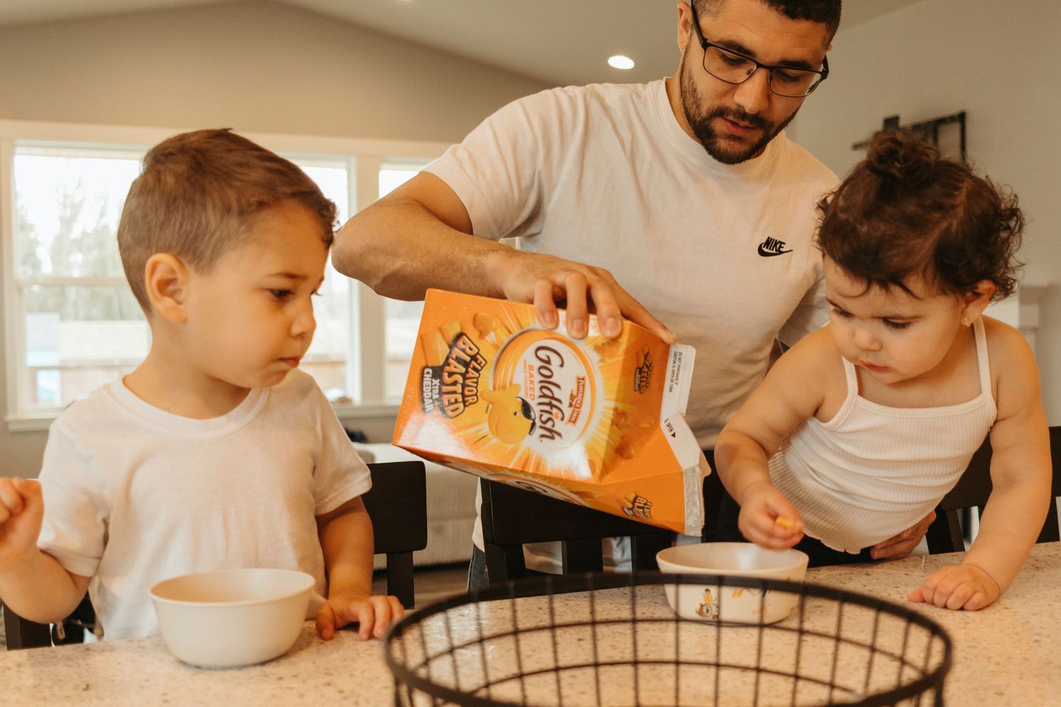 Two children watch attentively as food is poured into a large mixing bowl in a bright kitchen setting.