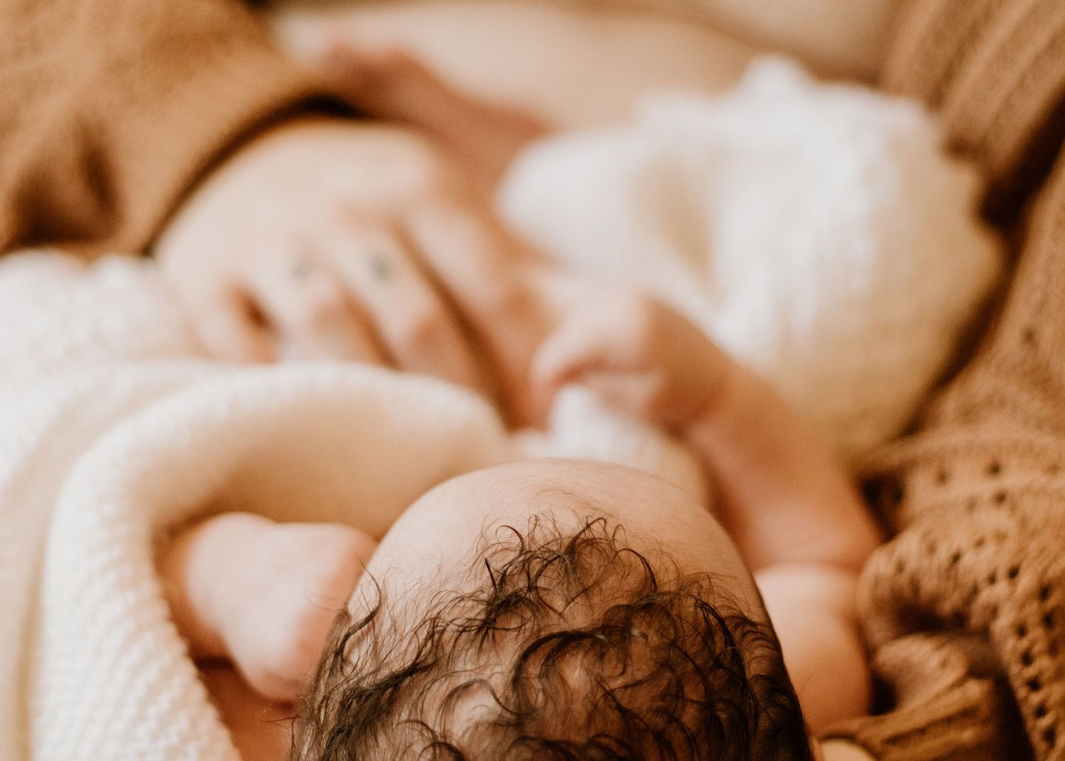 A baby sleeps peacefully on cozy cream-colored knit blankets in warm sepia-toned lighting.