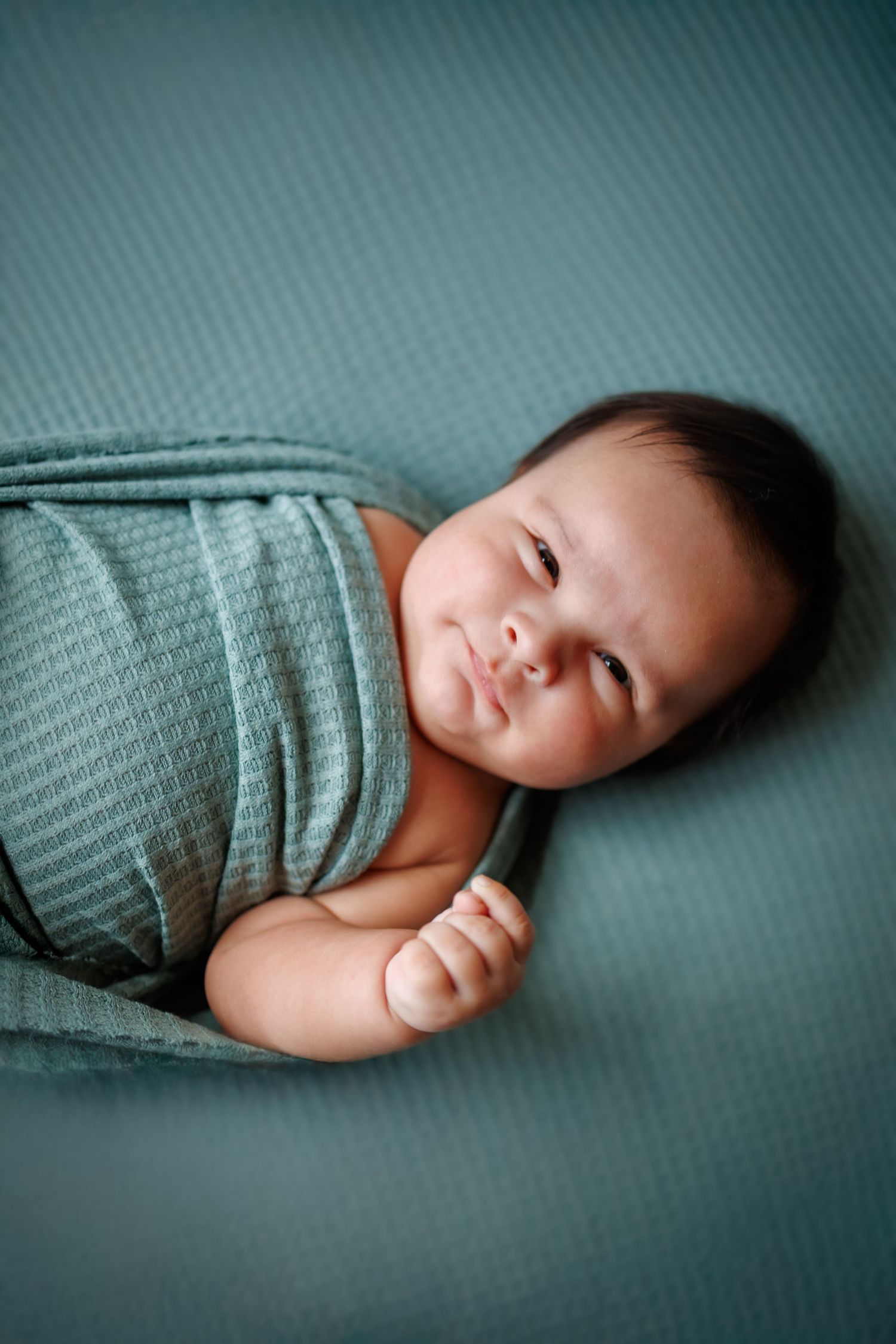 Sweet sleeping infant swaddled in soft teal blanket rests peacefully on teal surface in gentle lighting.