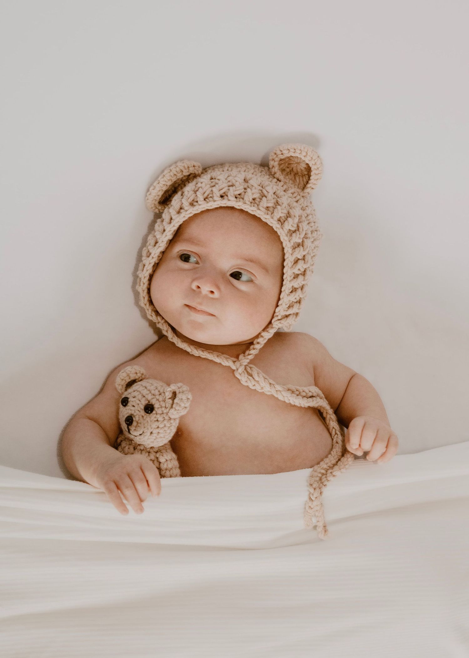 A baby wearing a crocheted beige bonnet lies on soft white bedding while holding a small teddy bear.