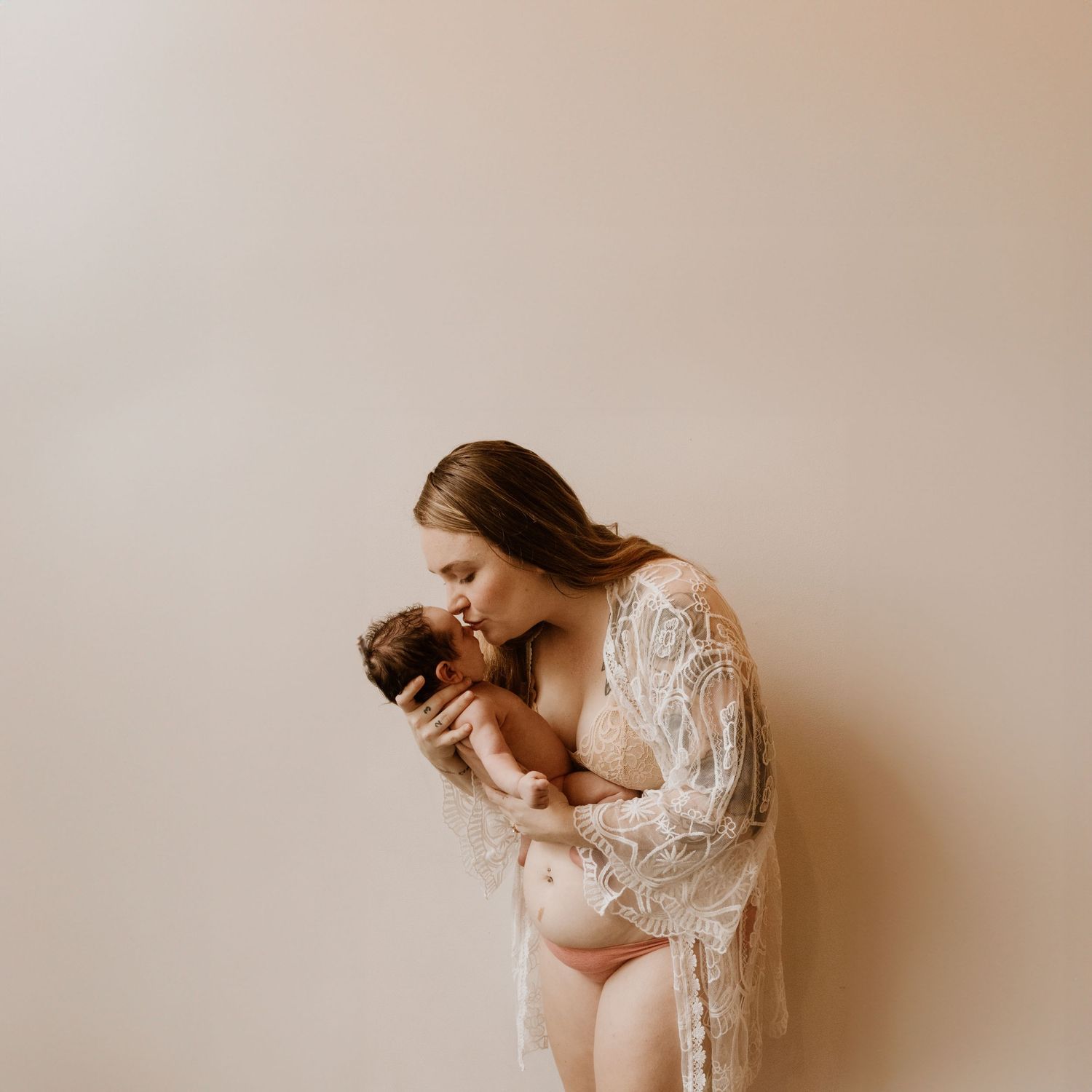 Intimate maternity boudoir photo featuring expecting mother in white lace robe holding puppy against neutral backdrop.