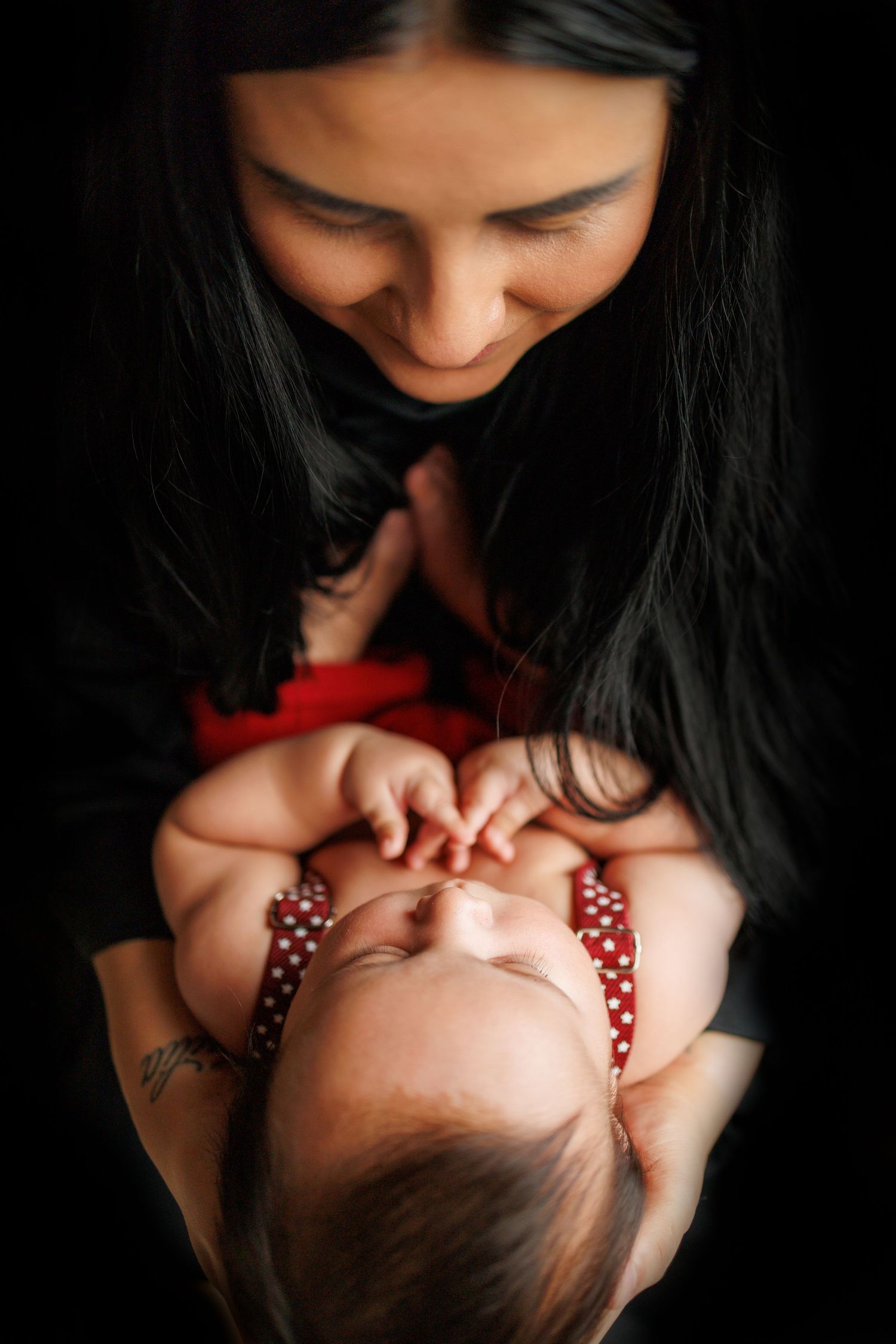 A mother in black clothing tenderly holds and kisses a small baby wearing a red and white polka dot outfit.