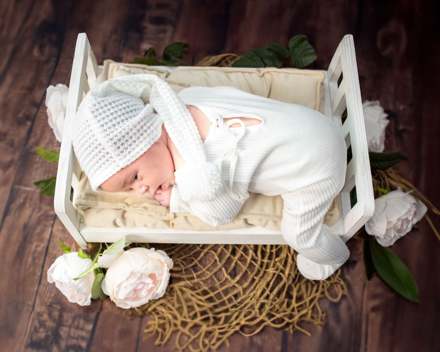 A newborn baby sleeping in a white wooden crate with pink roses and lace doily on dark wood floor.