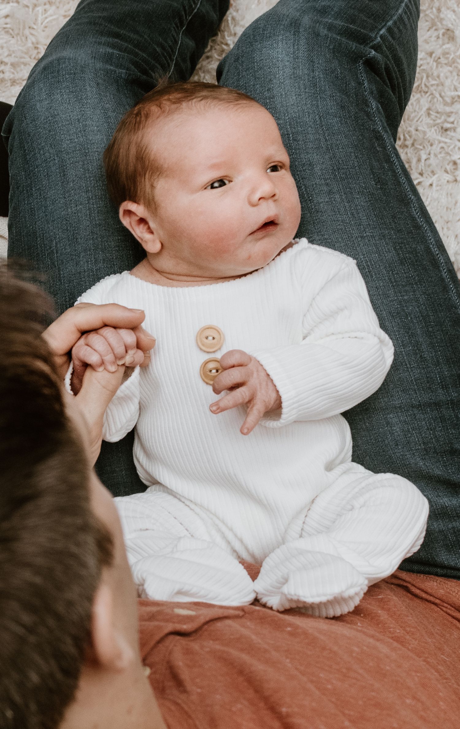 Tiny baby wearing white knit sweater rests on parents lap while being cradled in tender moment.