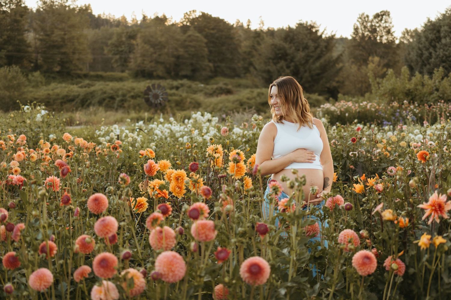 A person in a white dress stands in a colorful flower field at sunset with orange and pink dahlias blooming.