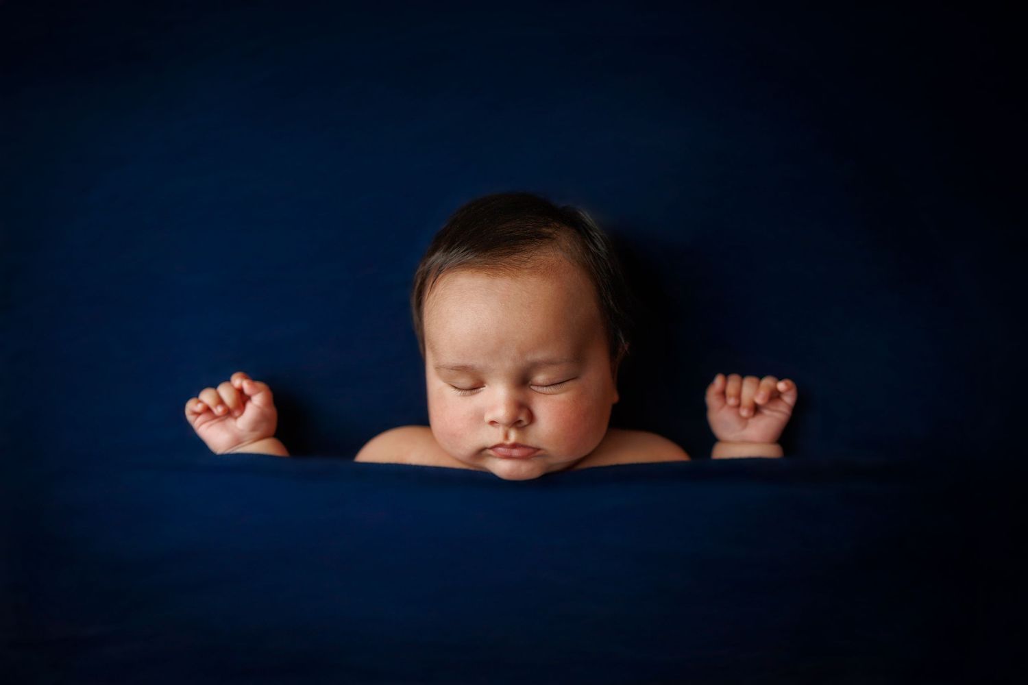 Sleeping baby in peaceful pose with raised fists against dark blue background in studio portrait.