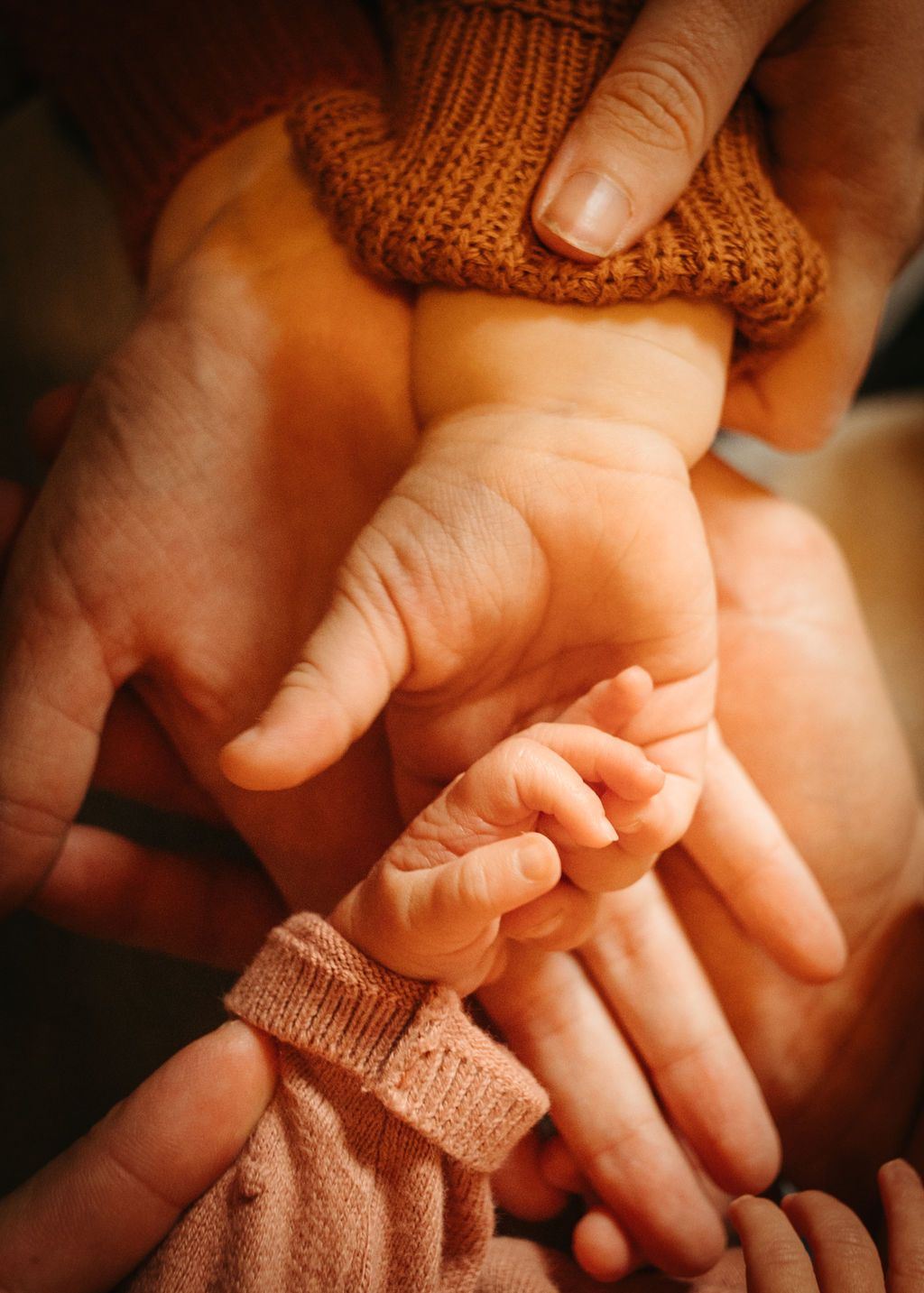 Close-up view of small hands gently holding a tiny baby's hand with knitted sleeve details in warm lighting.