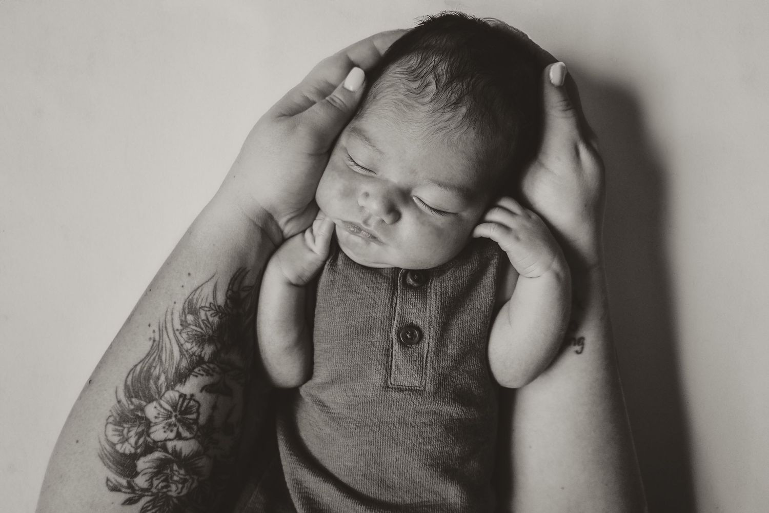 Black and white close-up of sleeping newborn being held by tattooed arms in gray onesie.