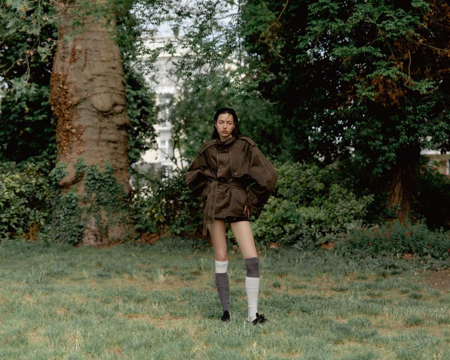 Woman in brown mini dress, knee-high socks, and black shoes standing on grass in a lush green park.