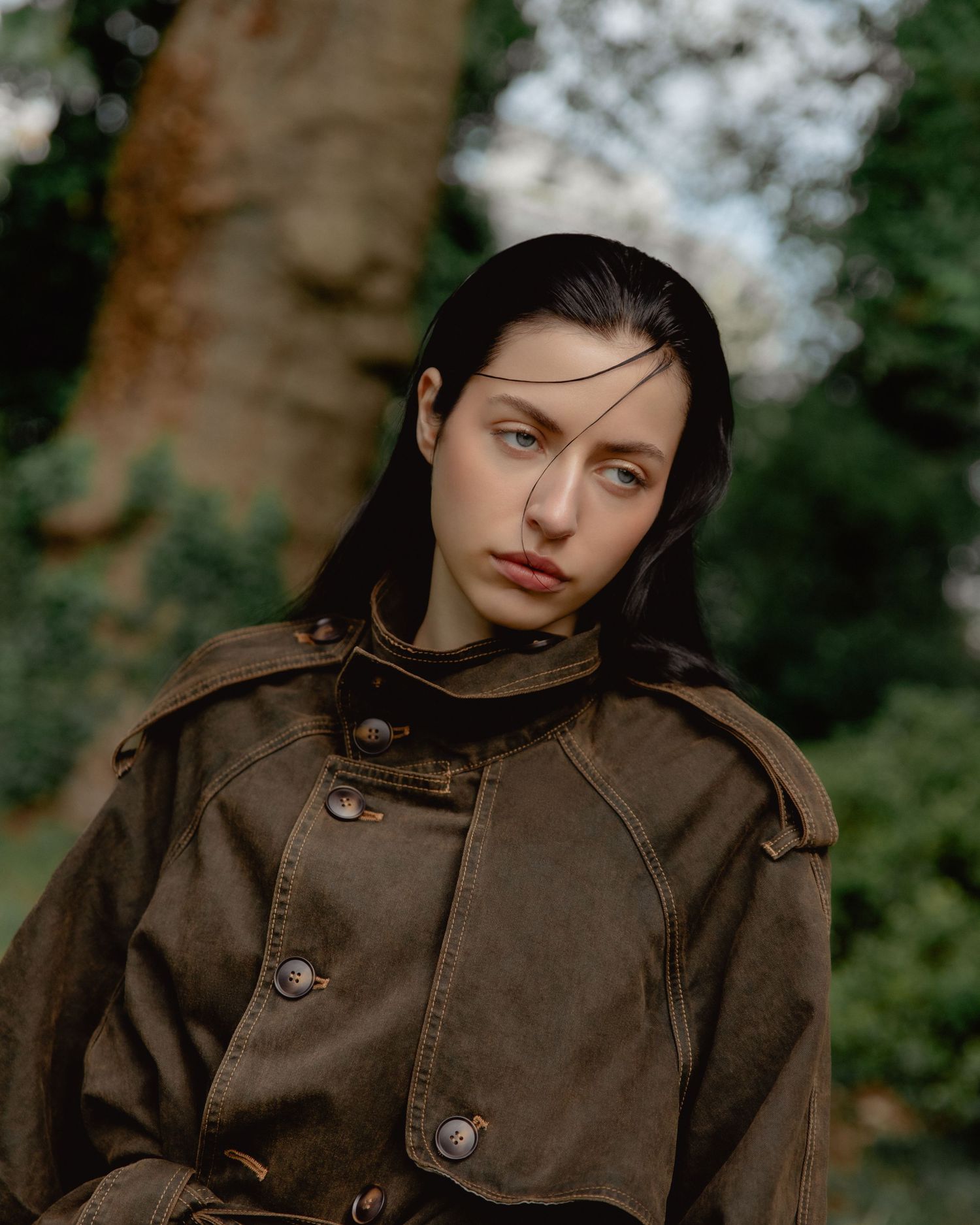 Young woman with dark hair wearing a brown trench coat poses outdoors near trees in a moody fashion portrait.