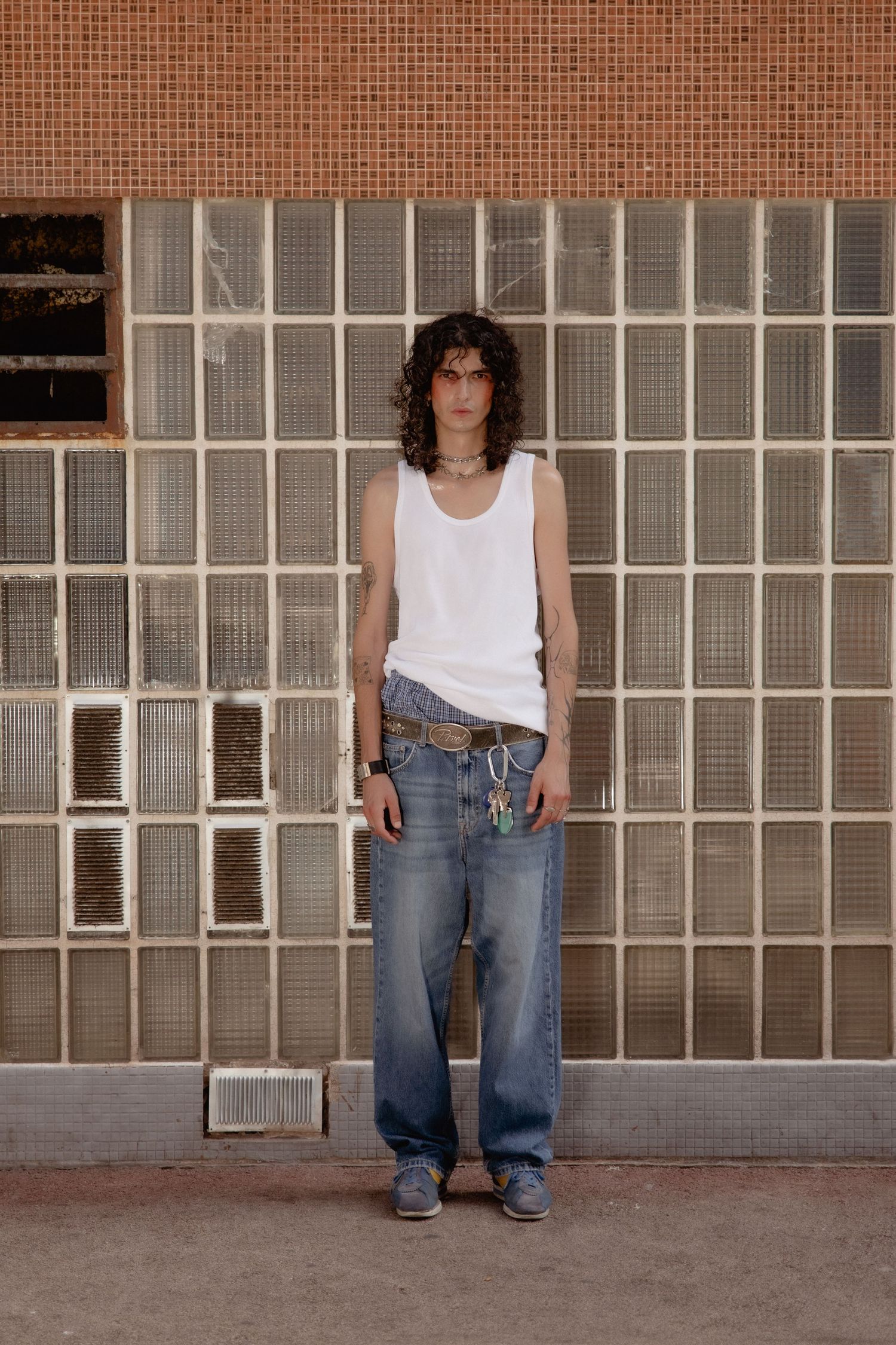 Person in a white tank top and baggy jeans poses against a brick wall with industrial windows.