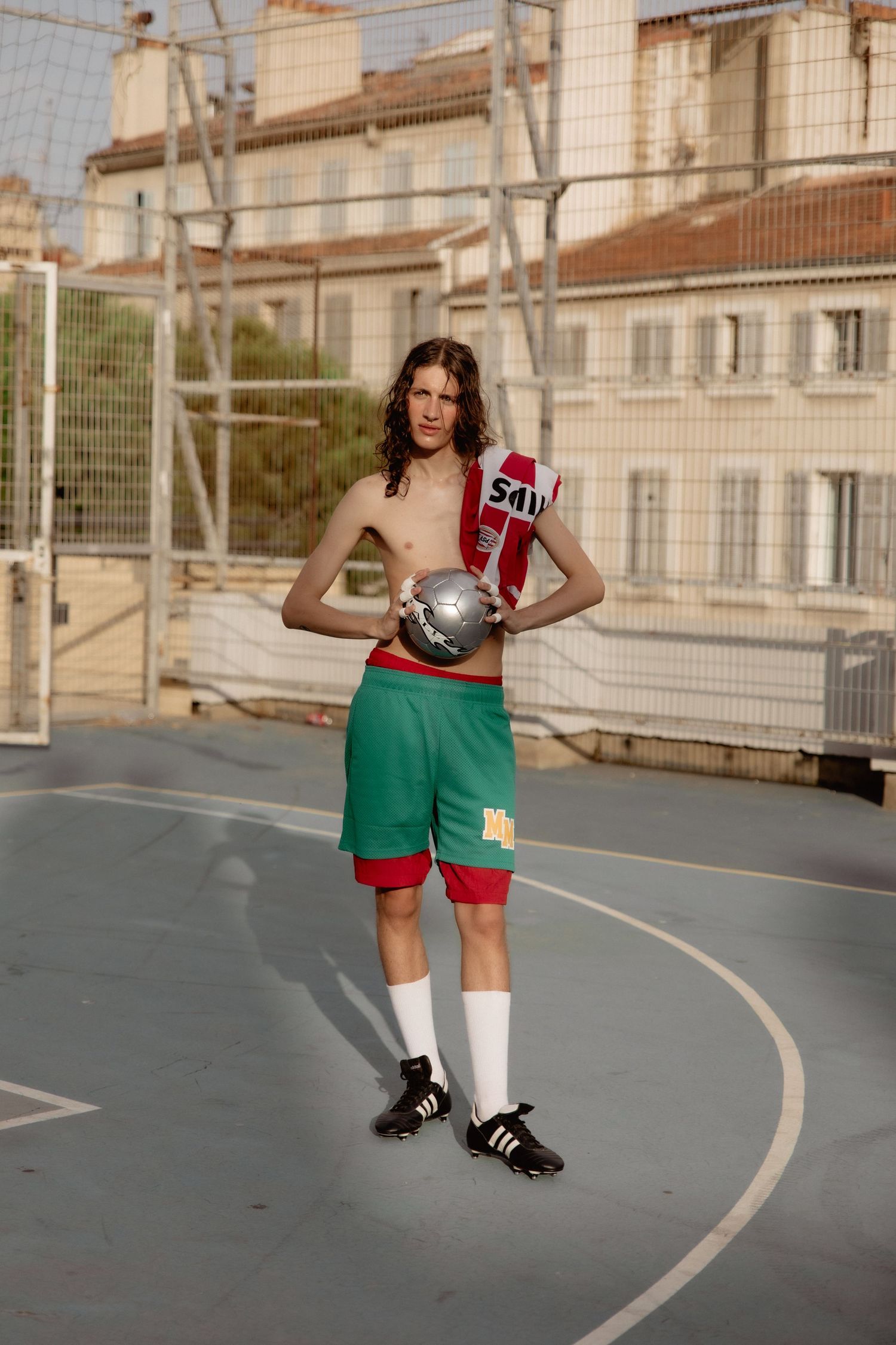 Athletic figure poses on outdoor basketball court wearing green and red shorts while holding a basketball and sports drink.