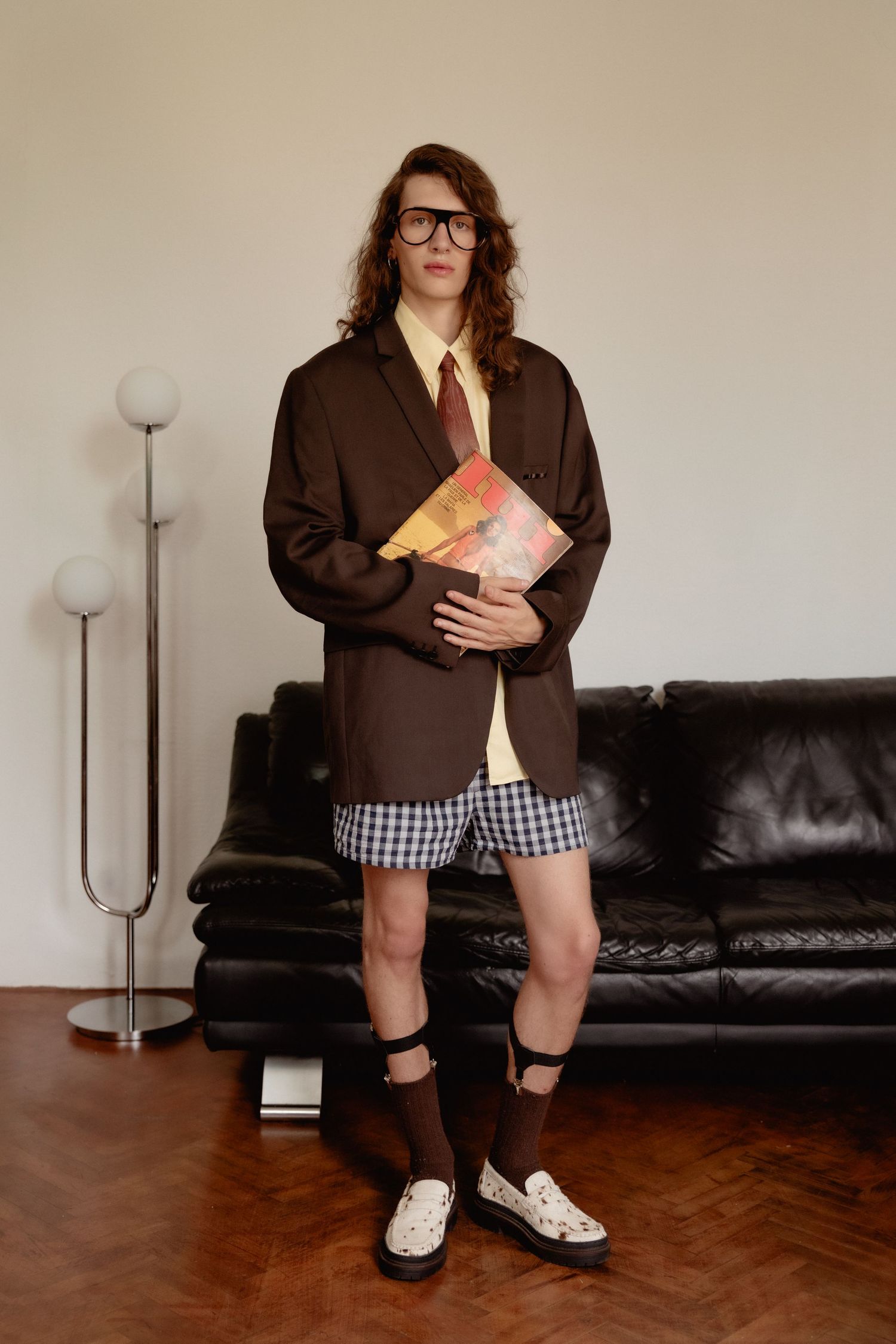 Person wearing brown blazer, checkered shorts, glasses and knee socks stands in living room holding books.