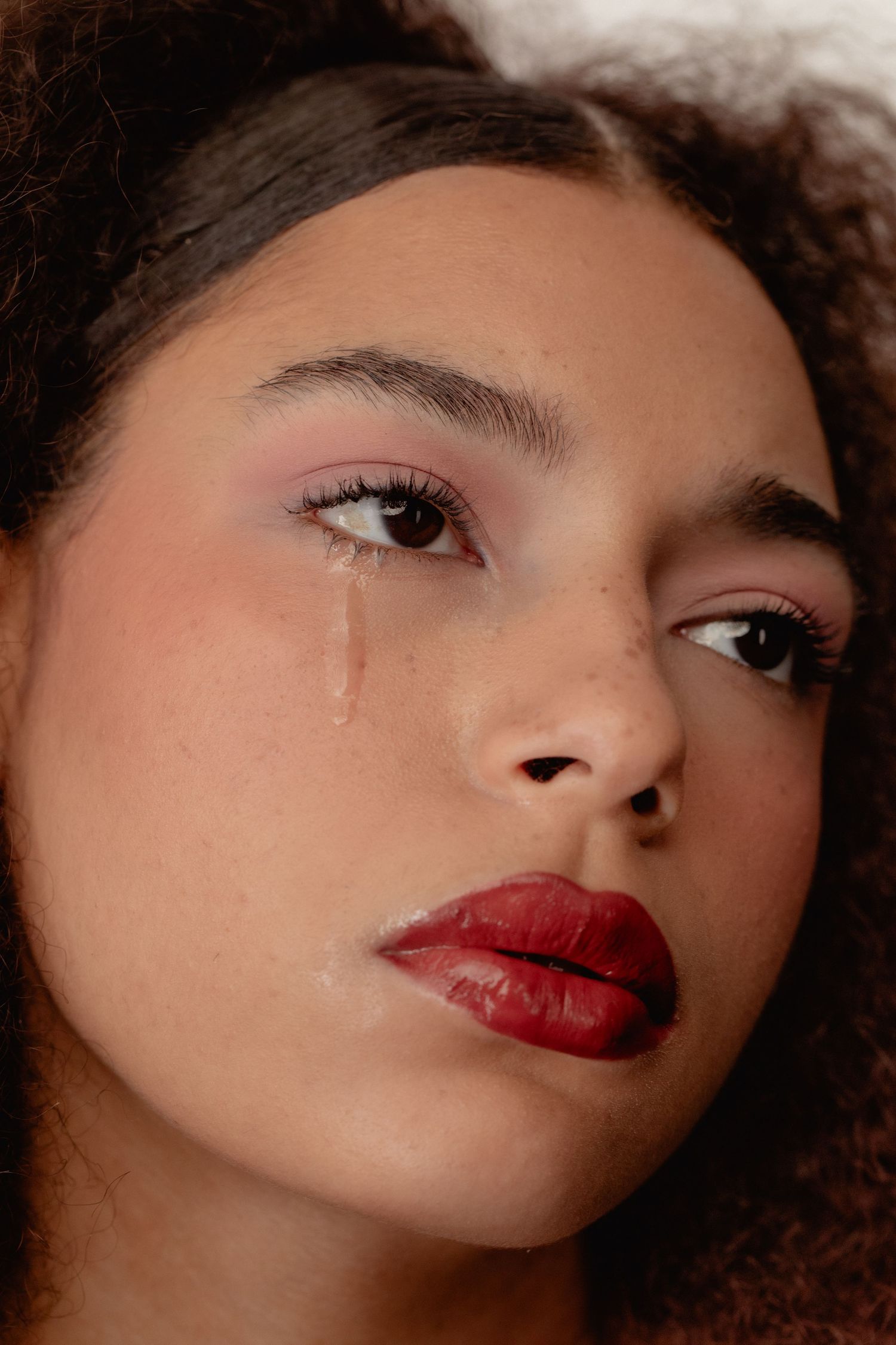 Close-up portrait of a young woman with red lips, pink eyeshadow, and a tear streaming down her cheek.