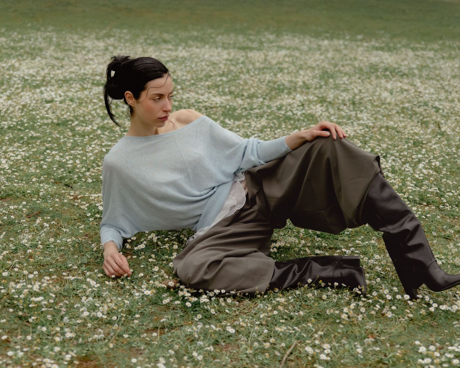 Woman in light blue sweater and gray trousers reclining in a field of white wildflowers.