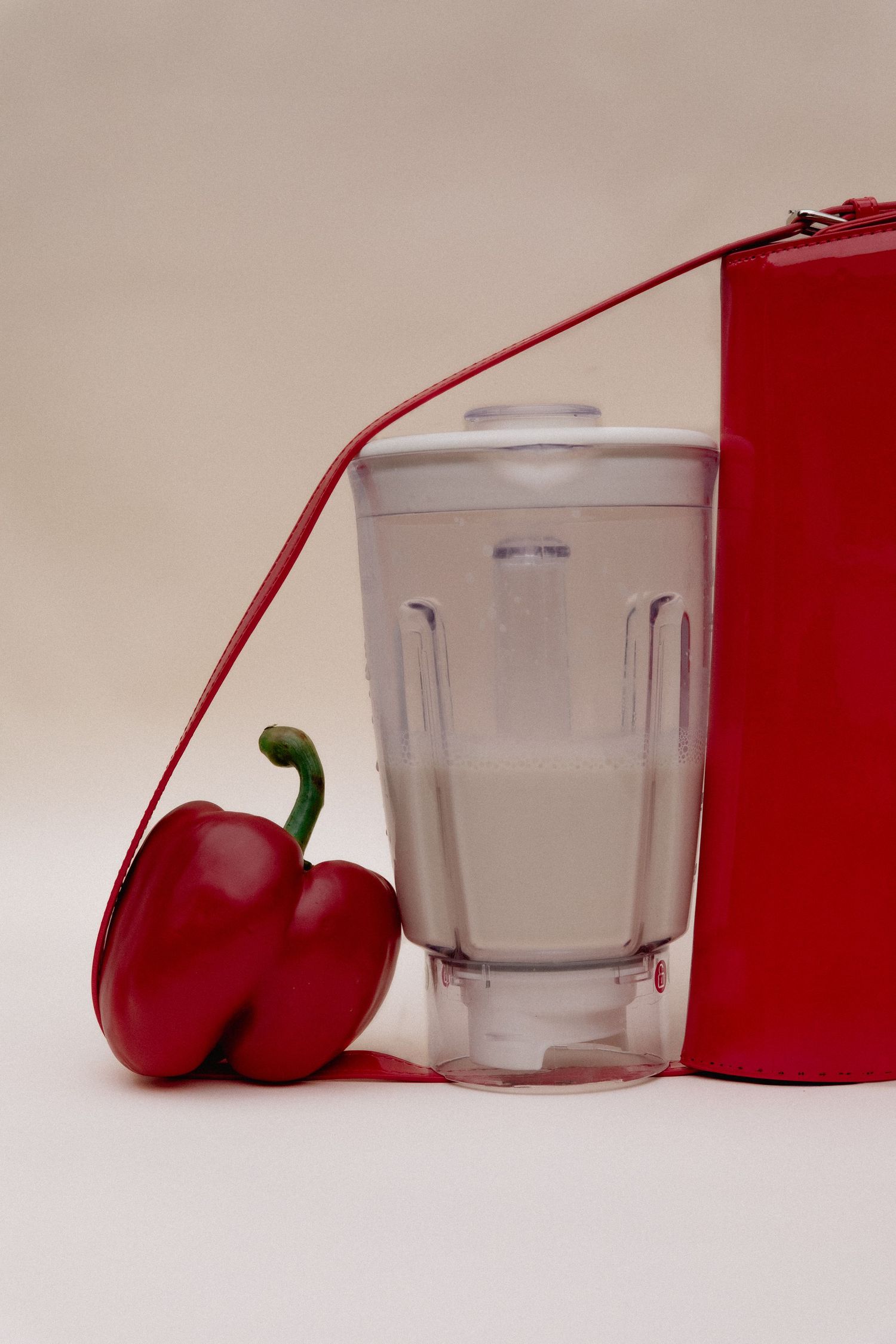 A glass of milk sits beside a fresh red bell pepper and red container against a neutral background.
