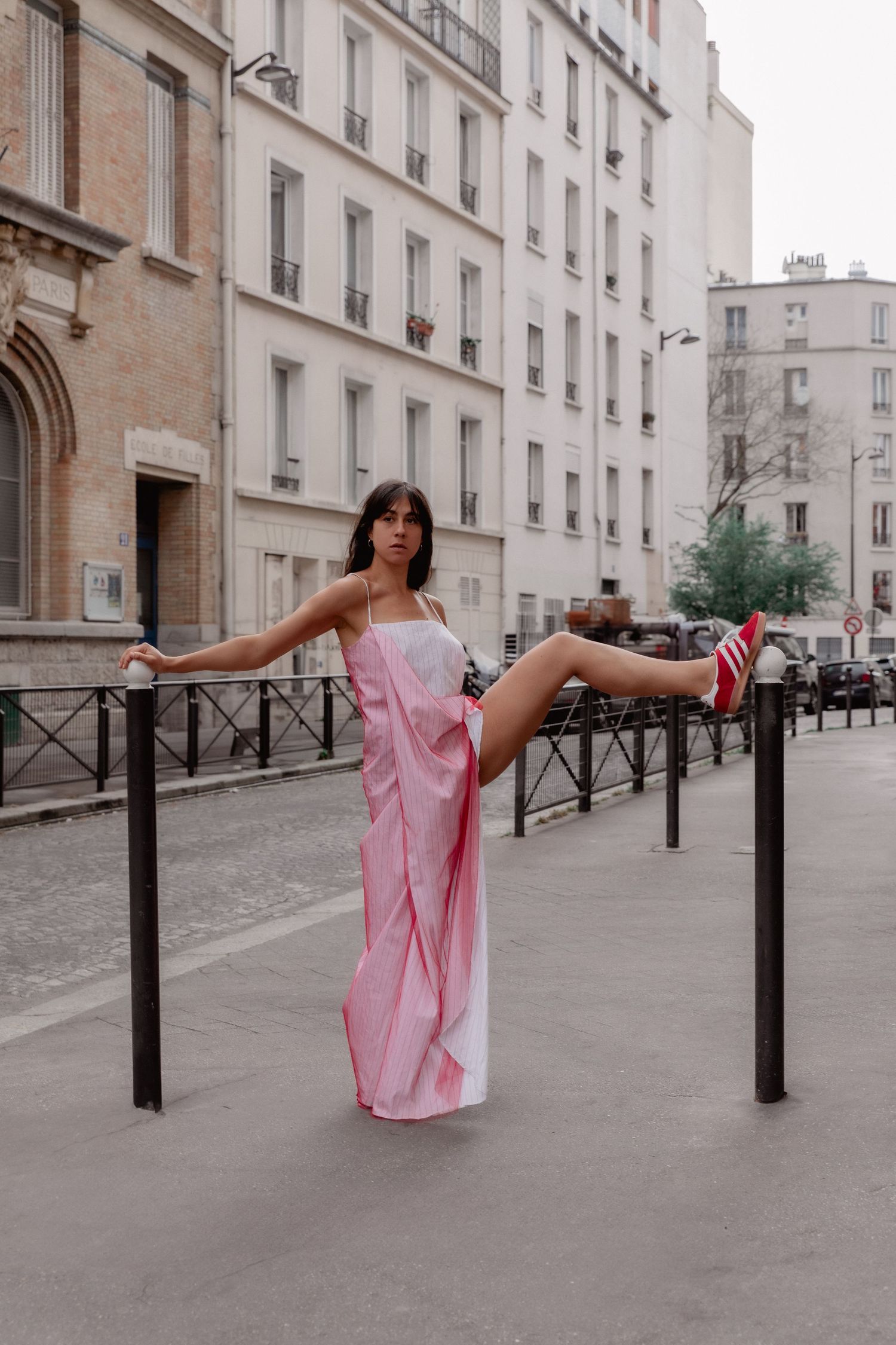 A person in a pink dress performs acrobatic moves on street barriers along a Parisian-style street with classic buildings.