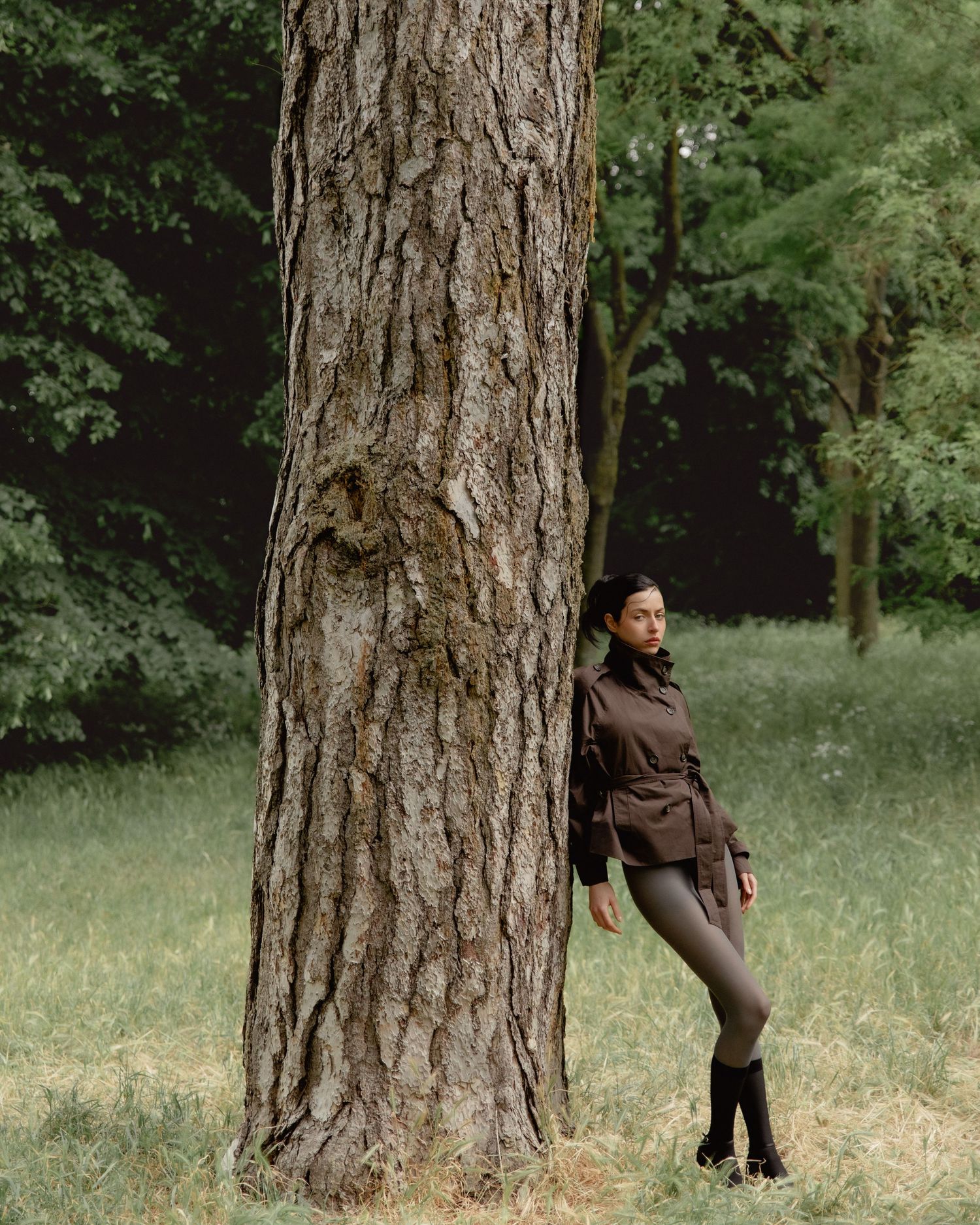 Woman in brown jacket and gray leggings leaning against large tree trunk in lush green forest park.
