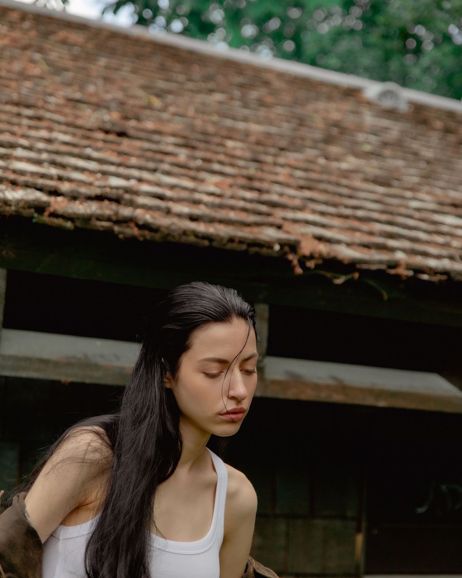 Young woman with long dark hair in a white tank top, eyes closed, standing before a rustic tiled roof building.