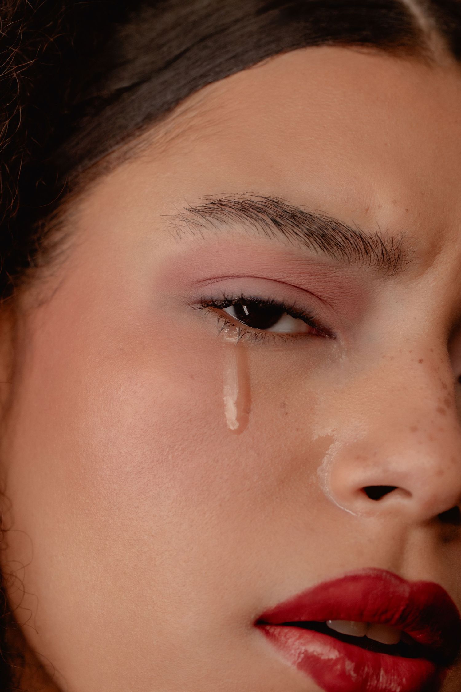 Close-up of a young woman with red lips and pink eyeshadow, a single tear streaming down her cheek.