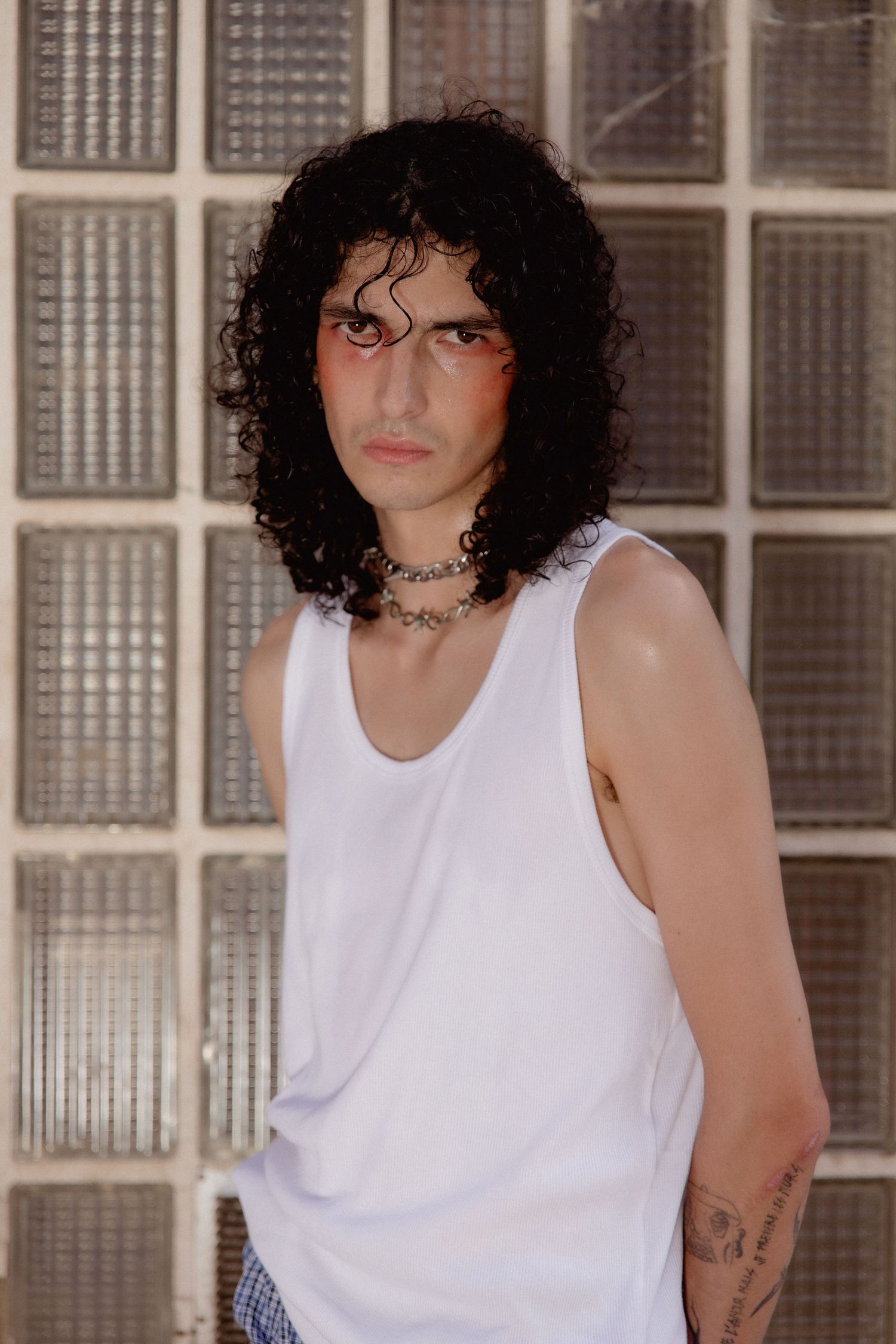 A person with curly dark hair and sunglasses wearing a white tank top poses against a window-lined wall.