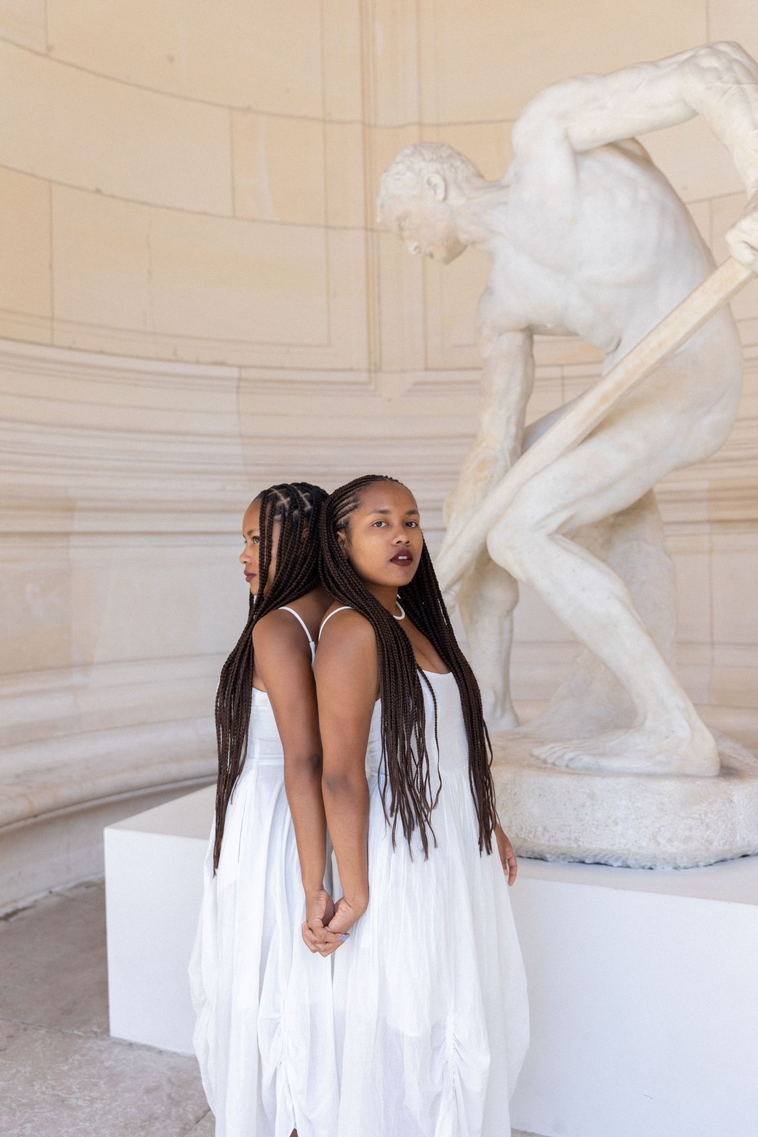 Woman in flowing white dress poses expressively next to classical marble sculpture.