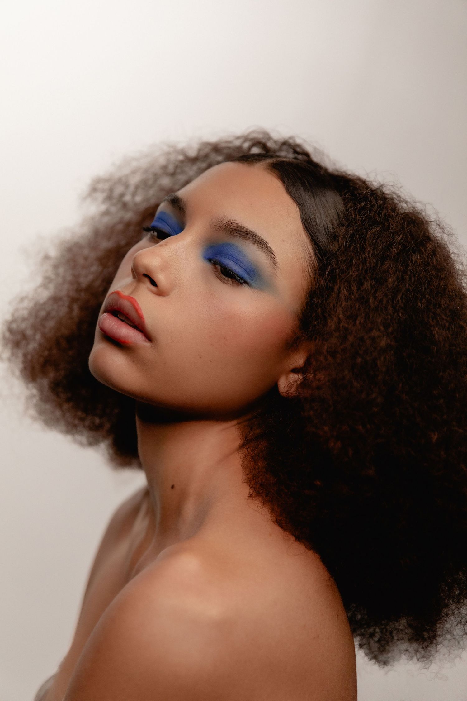 Young woman with curly hair and bold blue eyeshadow makeup poses against a light gray background.