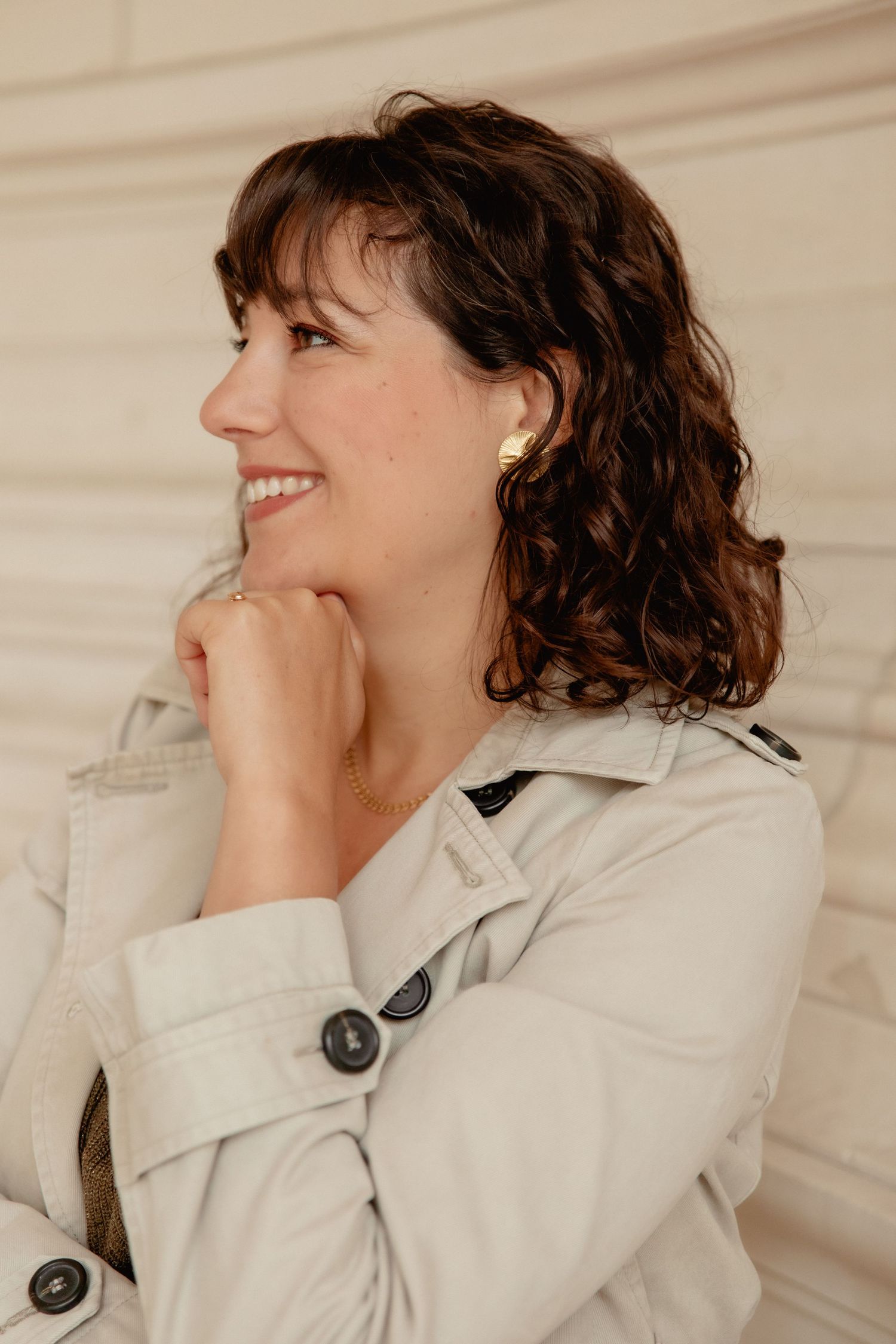 Side profile portrait of someone with curly dark hair wearing a beige trench coat smiling against a light background.