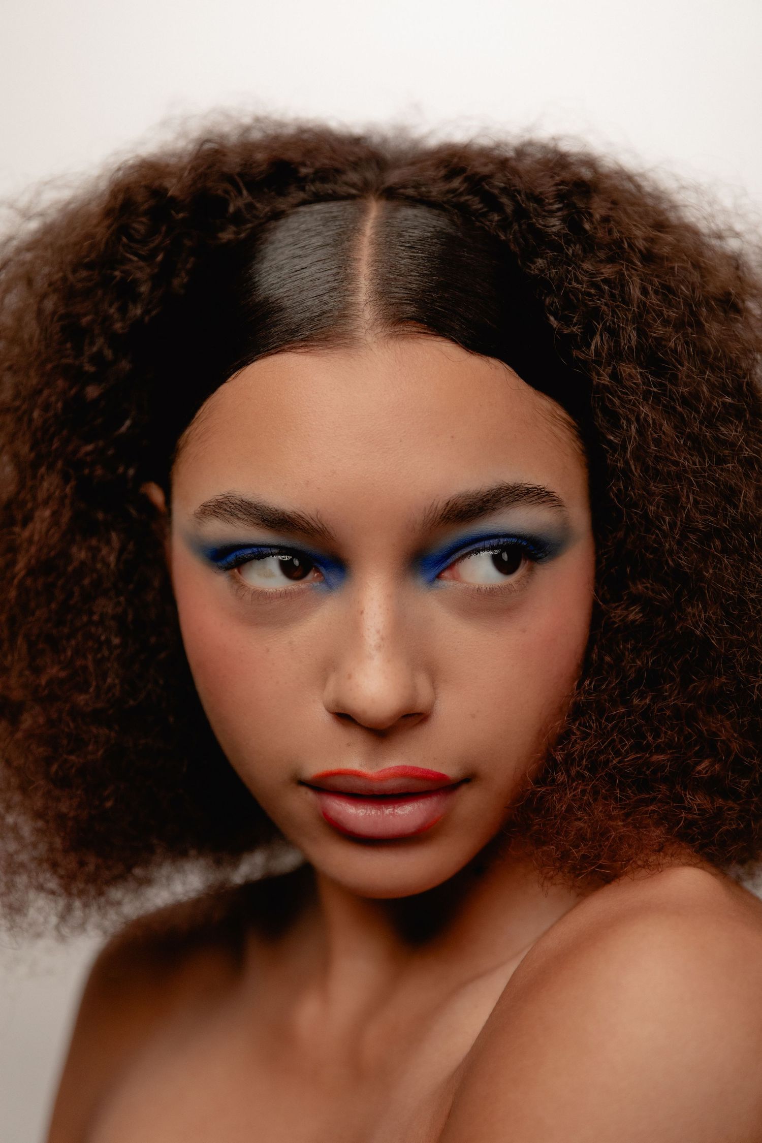 Close-up portrait of a woman with voluminous curly hair and bold blue graphic eye makeup and red lips.