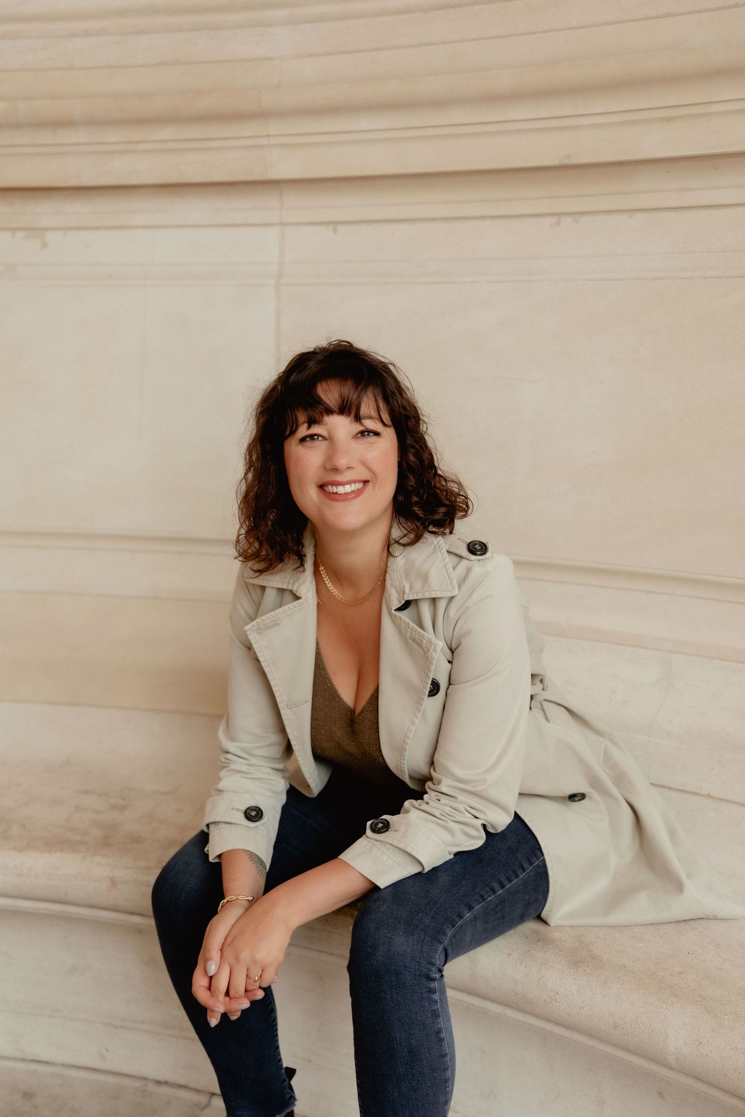 A person wearing a beige trench coat and jeans sits casually on light-colored steps in a minimalist setting.