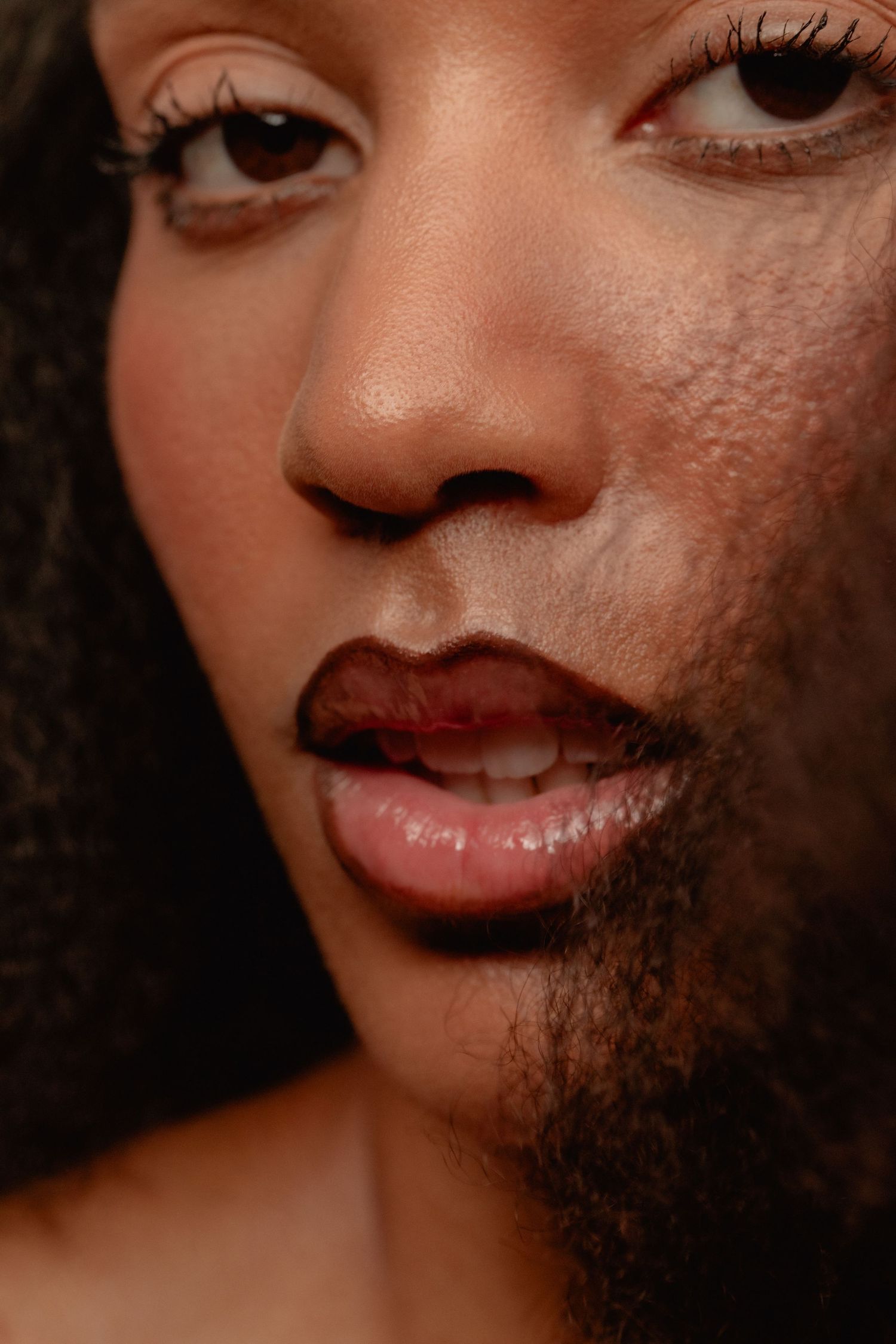 Close-up portrait of a woman with dark lipstick, long lashes, and natural curly hair against a dark background.