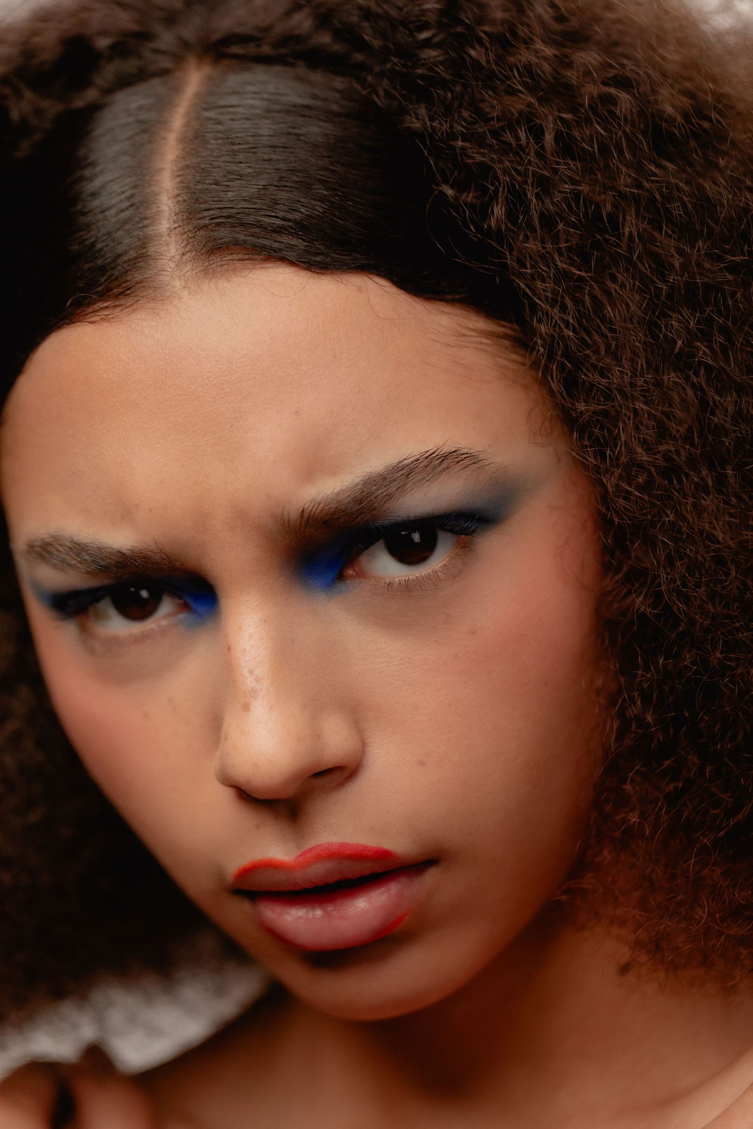 Close-up portrait of a young woman with curly hair, bold blue eyeliner, and red lips wearing dramatic makeup.